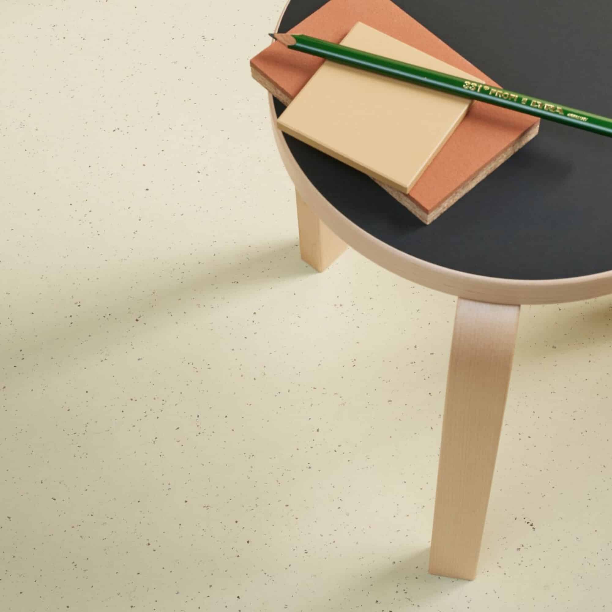Ein runder Tisch mit einem schwarzen Tischblatt und hellen Holzbeinen, darauf liegen ein grüner Bleistift, ein beige-farbenes Notizblatt und ein braunes Stück Papier. Der Boden ist in einem hellen Farbton mit kleinen, bunten Punkten.