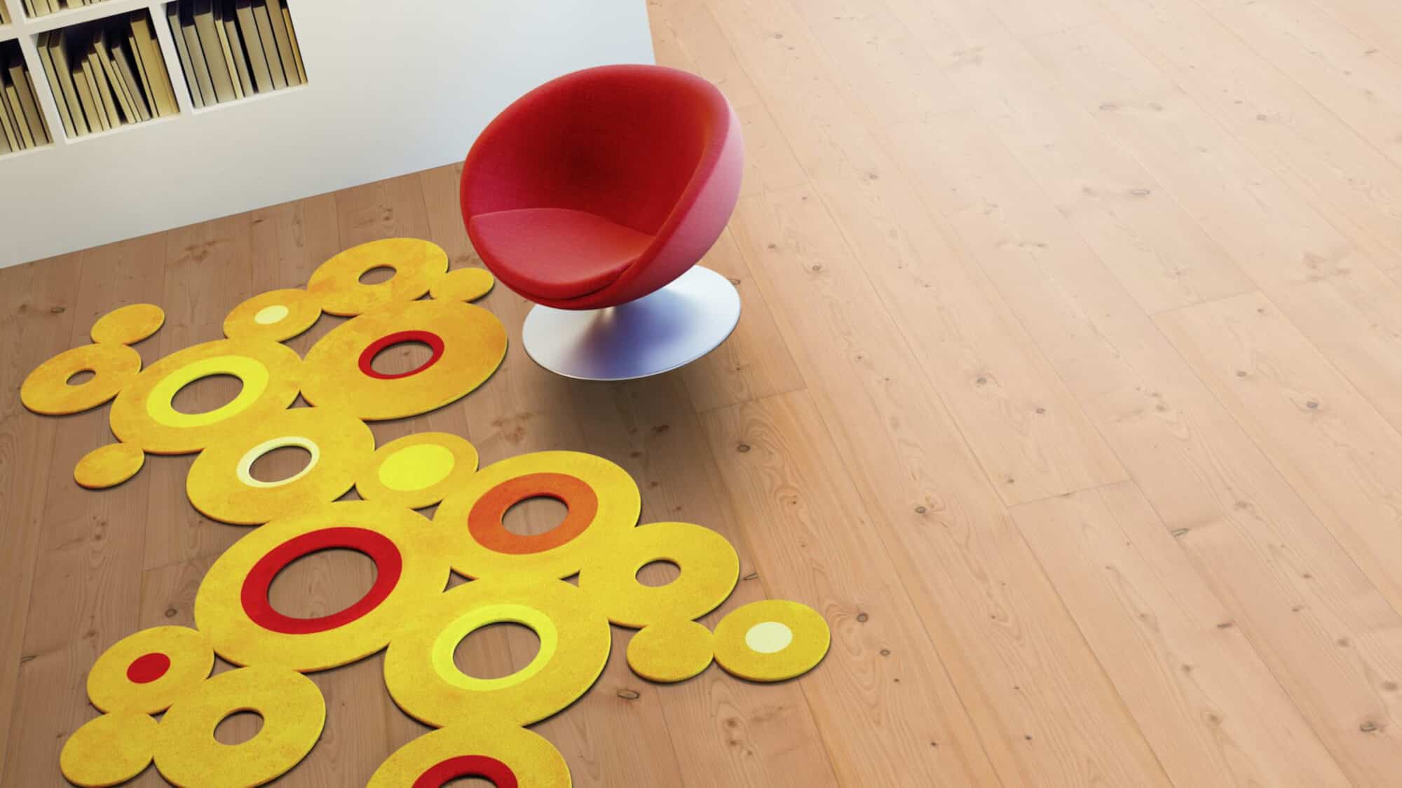 Ein roter Sessel steht auf einem bunten, kreisförmigen Teppich in Gelb- und Rottönen. Der Boden ist aus hellem Holz, und im Hintergrund sind Bücherregale sichtbar.