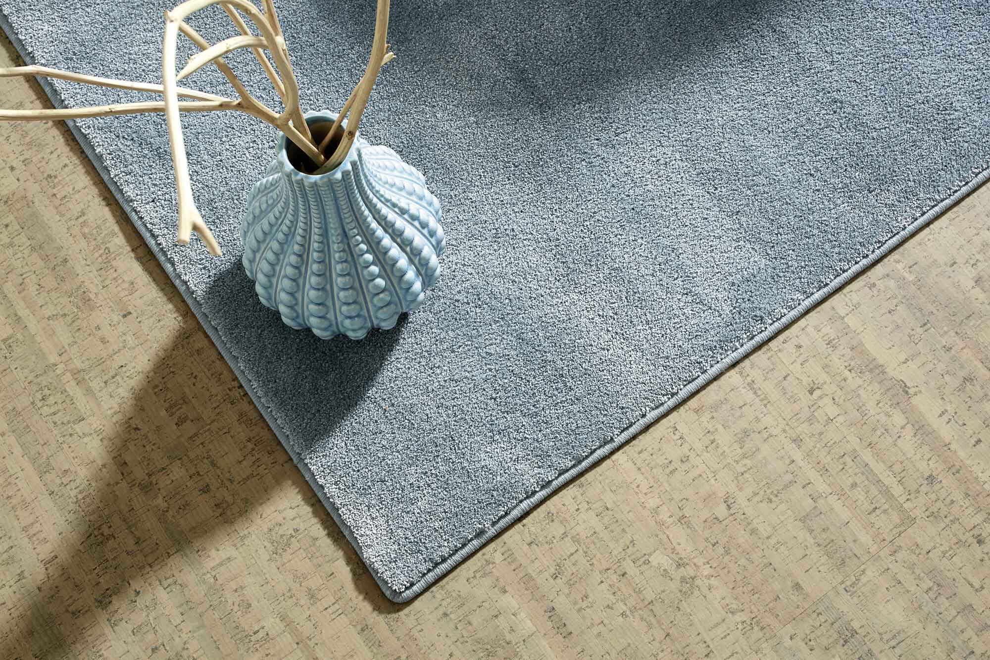 Ein blauer Teppich liegt auf einem hellen, strukturierten Boden. Darauf steht eine elegante, muschelförmige Vase mit trockenen Zweigen.