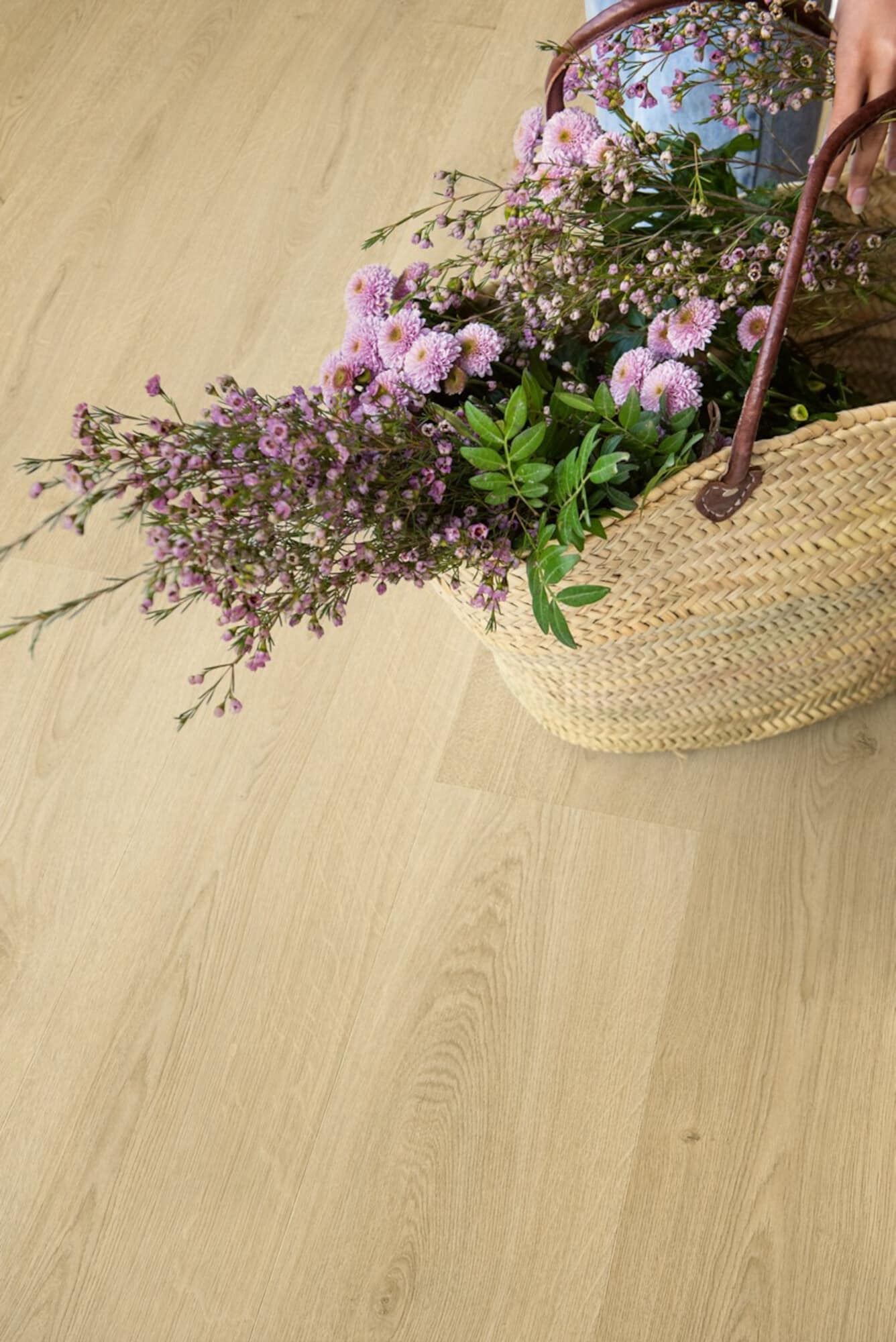Ein geflochtener Korb mit bunten Blumen, darunter zarte lila Blüten und grüne Blätter, steht auf einem hellen Holzboden. Eine Hand hält den Korb, während die Blumen herausragen.