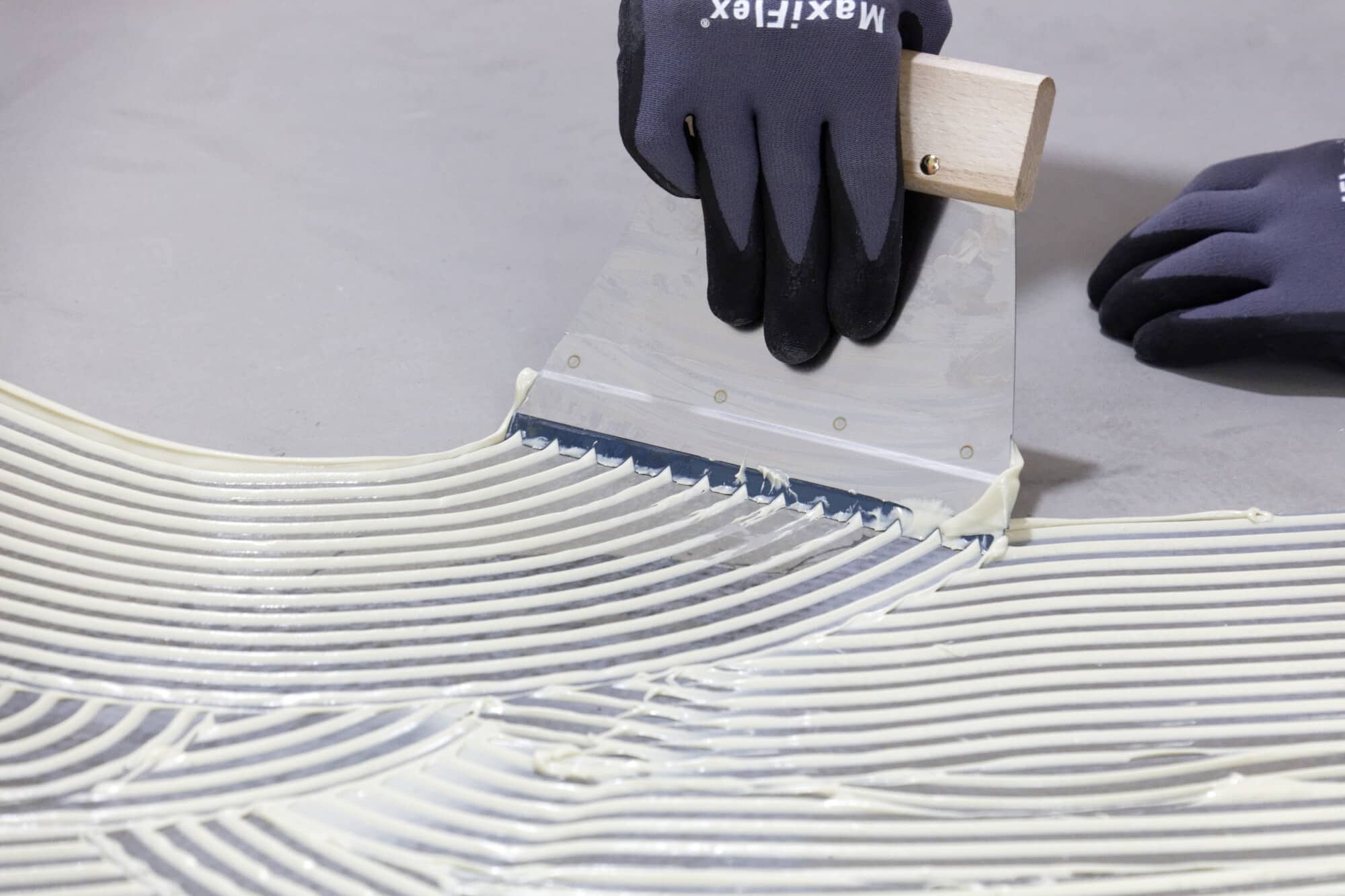Eine Hand trägt mit einer Kelle einen weißen Kleber auf eine glatte Oberfläche auf, wobei gleichmäßige Linien entstehen. Die Person trägt schwarze Handschuhe und nutzt eine Kelle mit einem Holzgriff.
