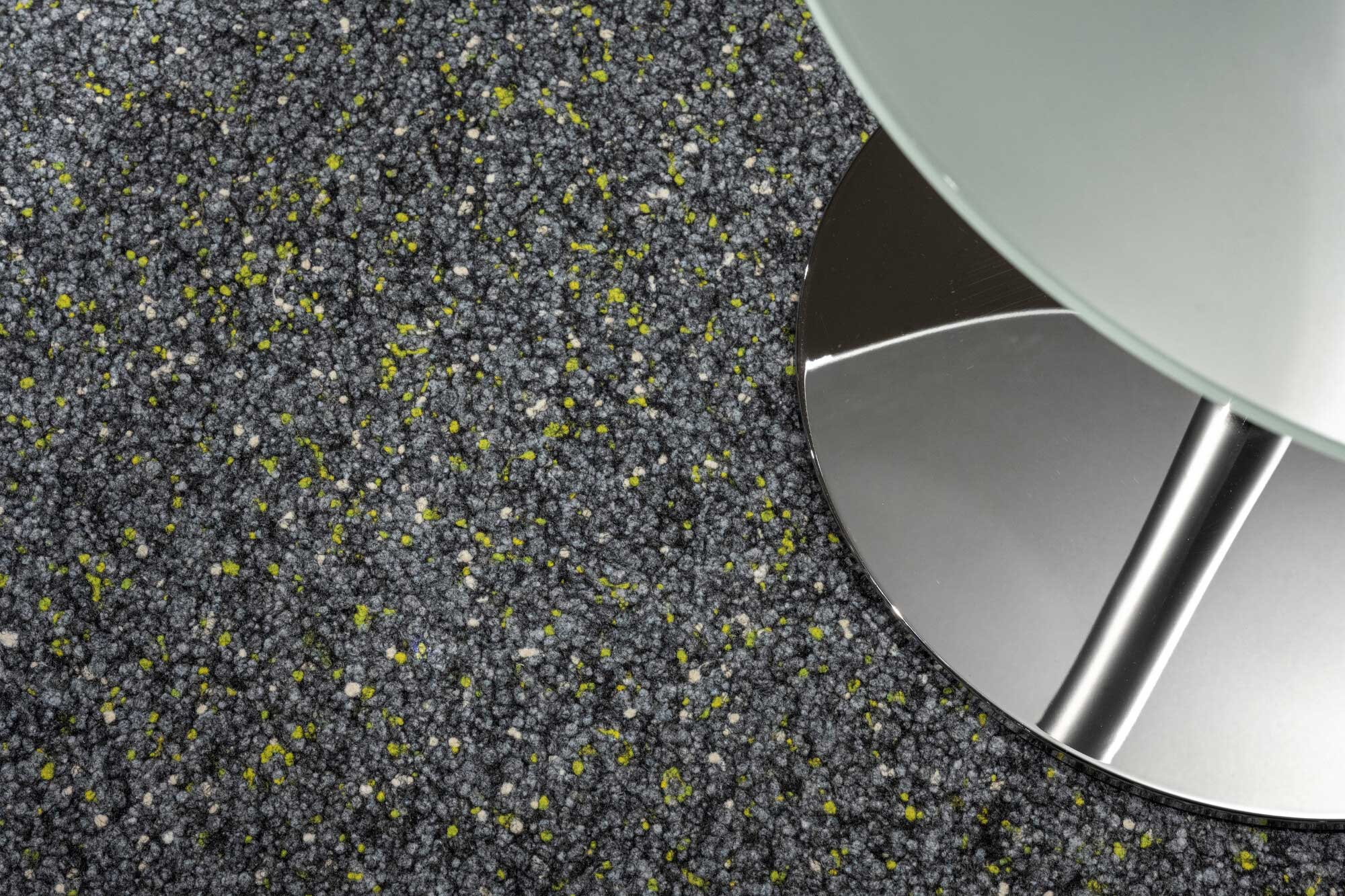 Ein grauer Teppich mit einem strukturierten Muster, durchzogen von gelben und weißen Farbakzenten, im Vordergrund steht eine Tischplatte aus Glas mit einem glänzenden, runden Metallfuß.