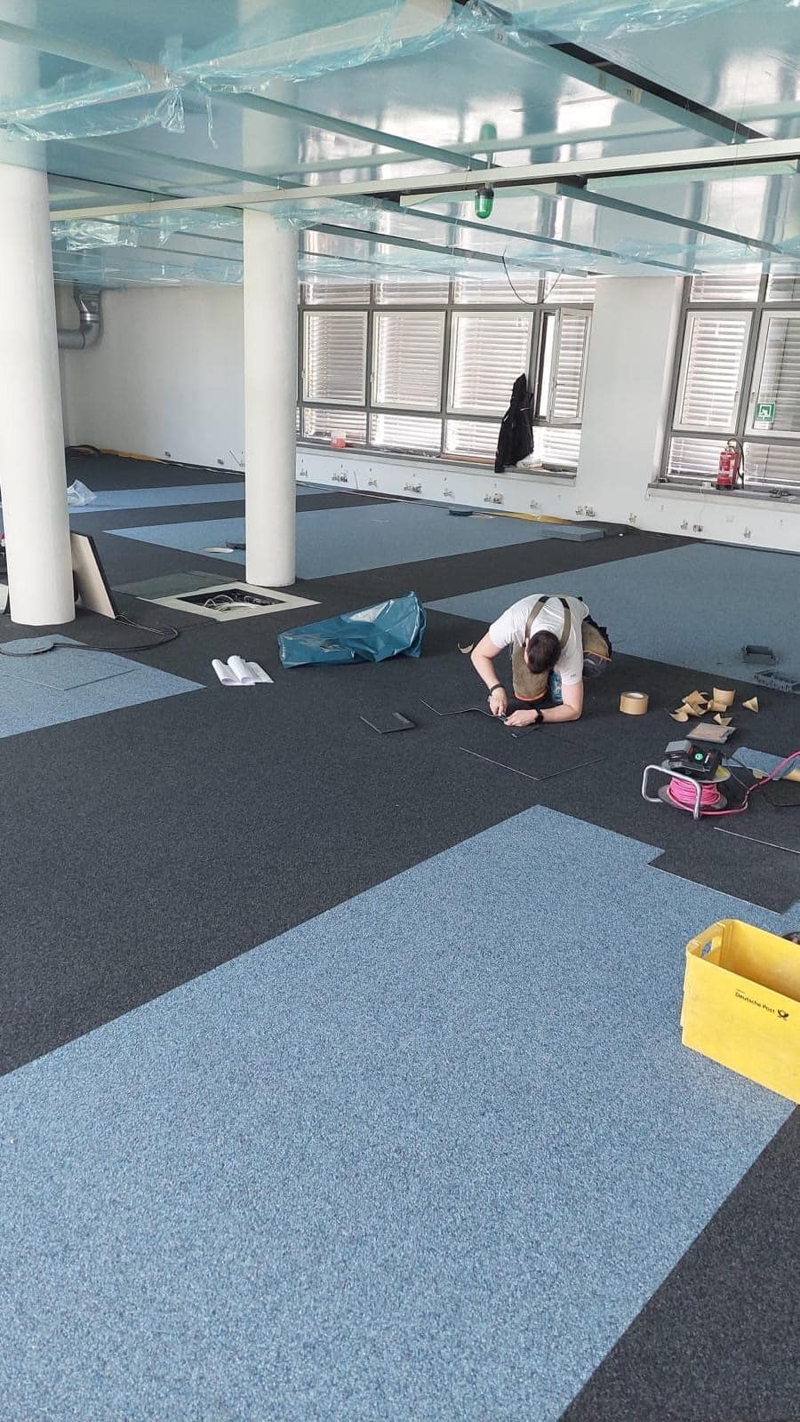In einem modernen Büro wird ein neuer Teppichboden verlegt. Ein Handwerker kniet auf dem Boden und arbeitet an den Teppichfliesen, während verschiedene Materialien und Werkzeuge um ihn herum liegen. Die Wände sind hell und mit großen Fenstern ausgestattet, die für viel Tageslicht sorgen.