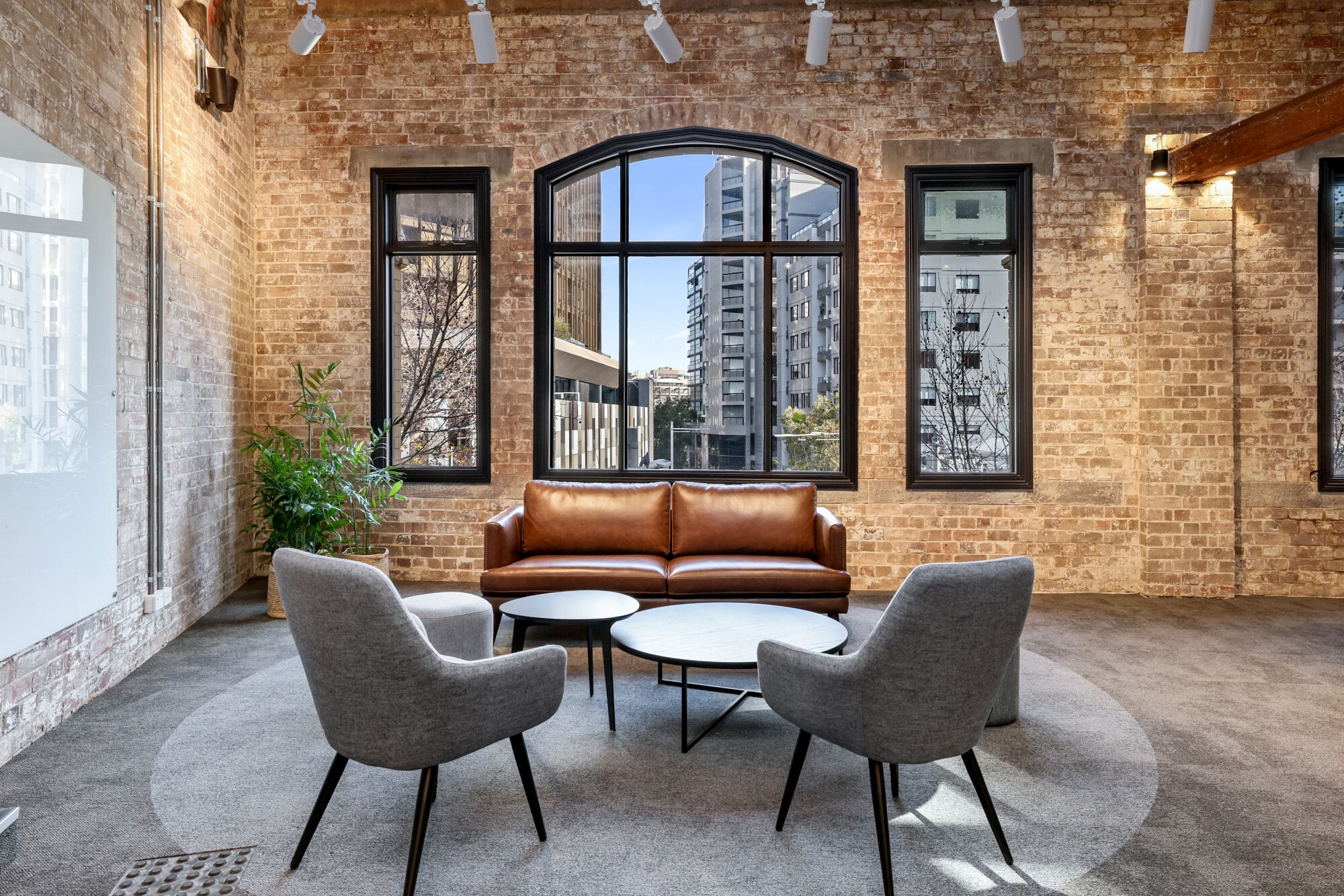 Ein modernes Wohnzimmer mit einer braunen Ledercouch, zwei grauen Sessel und einem runden Tisch. Große Fenster lassen viel Tageslicht herein und bieten einen Blick auf die Stadt. Die Wände sind aus rotem Ziegelstein, und der Boden ist mit grauem Teppich ausgelegt. Eine Pflanze steht in der Ecke und sorgt für ein angenehmes Ambiente.