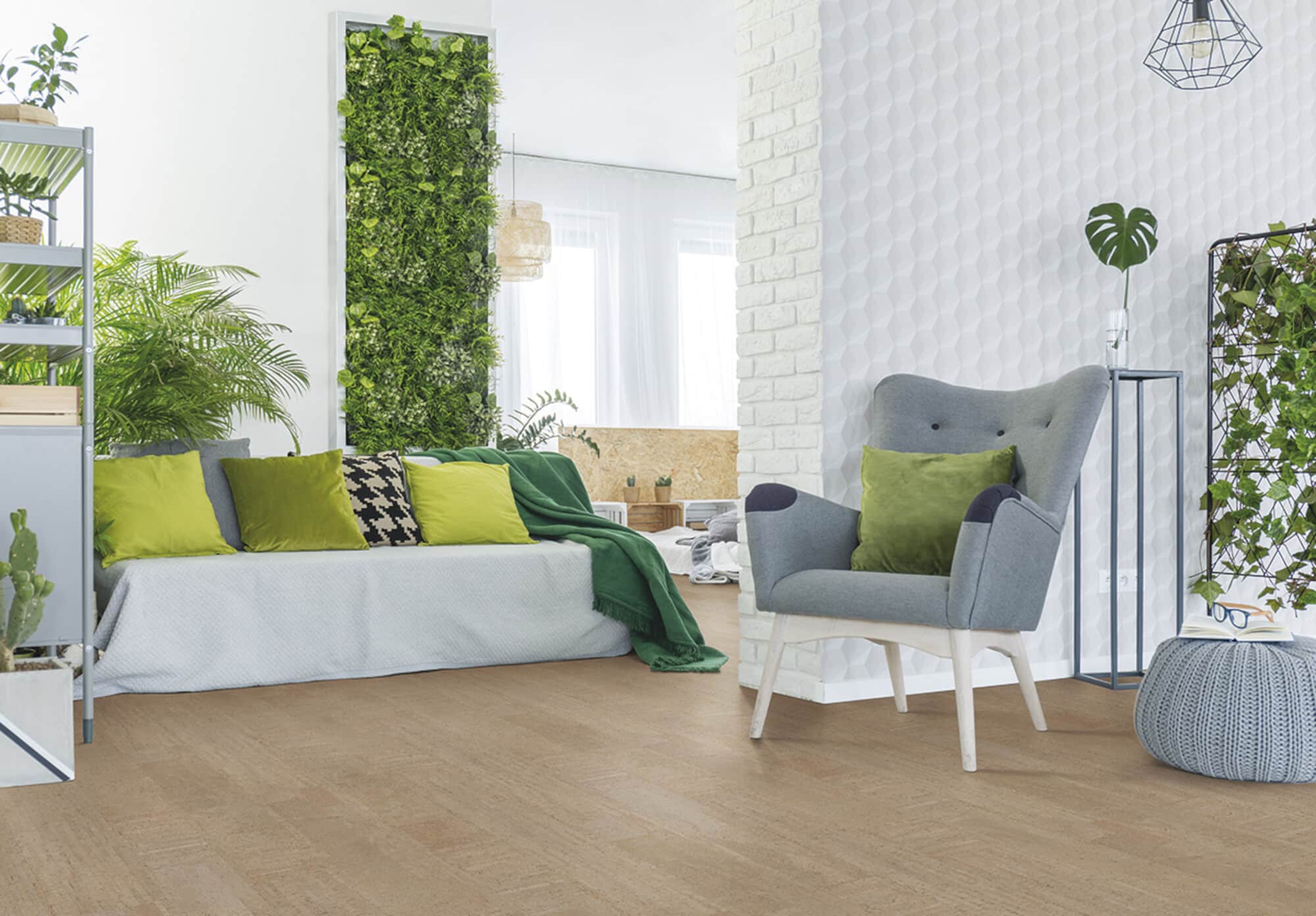 Gemütliches Wohnzimmer mit einer hellen Couch, bunten Kissen und einer grünen Decke. Im Hintergrund befindet sich eine grüne Pflanzenwand und ein grauer Sessel. Der Raum hat helle Wände und einen warmen Holzboden. Ein Tisch mit Pflanzen und eine moderne Lampe ergänzen die Einrichtung.