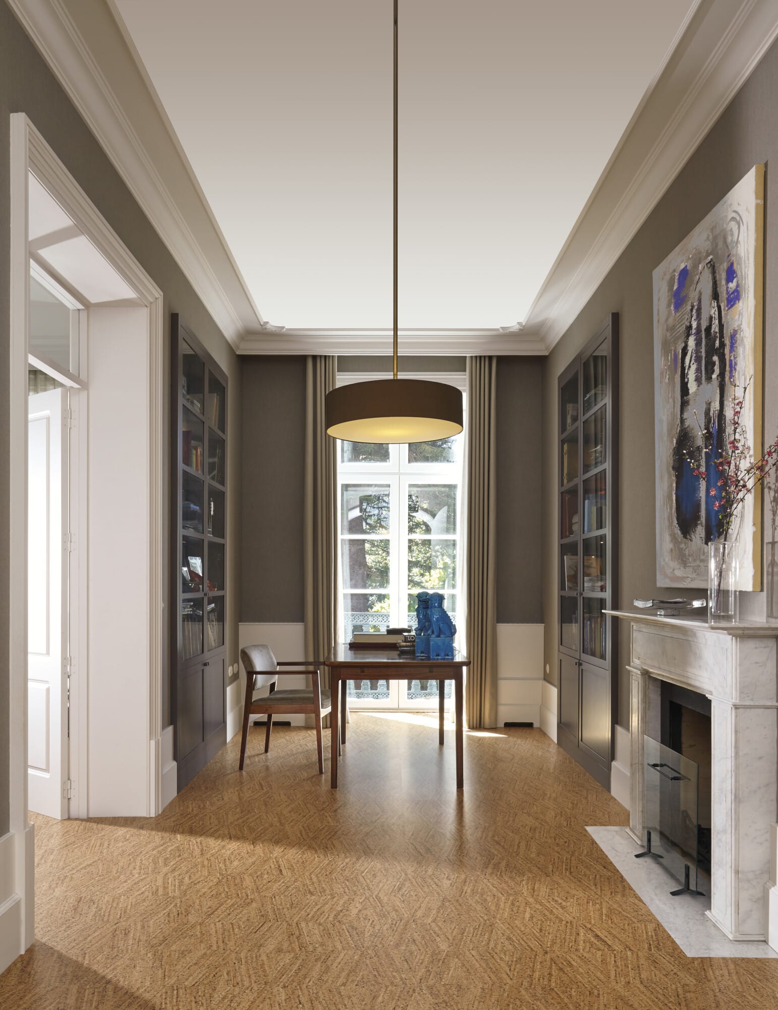 Ein elegantes Arbeitszimmer mit einem Holzfußboden, einem großen Fenster mit Vorhängen, einem schlichten Tisch und einem Stuhl. An den Wänden stehen Bücherregale, und ein moderner Kronleuchter hängt von der Decke. An einer Wand befindet sich ein Kamin mit einem Marmorrahmen und einem Kunstwerk daneben.