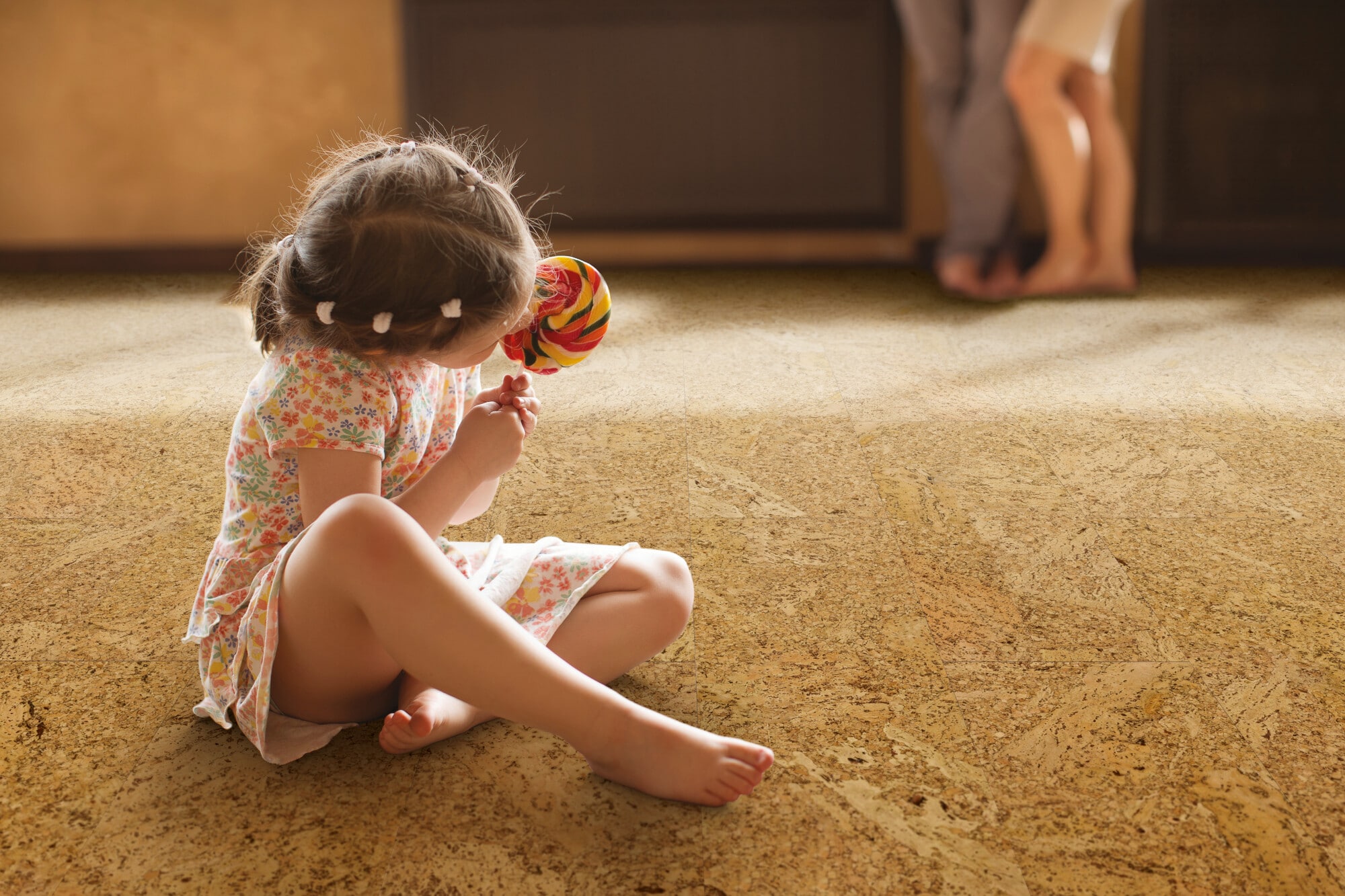Ein kleines Mädchen sitzt auf dem Boden und hält einen bunten Lutscher in der Hand. Sie trägt ein blumiges Kleid und hat ihre Beine angezogen. Im Hintergrund sind zwei Erwachsene zu sehen, die miteinander sprechen. Der Boden ist aus hellem Material und die Atmosphäre wirkt warm und einladend.