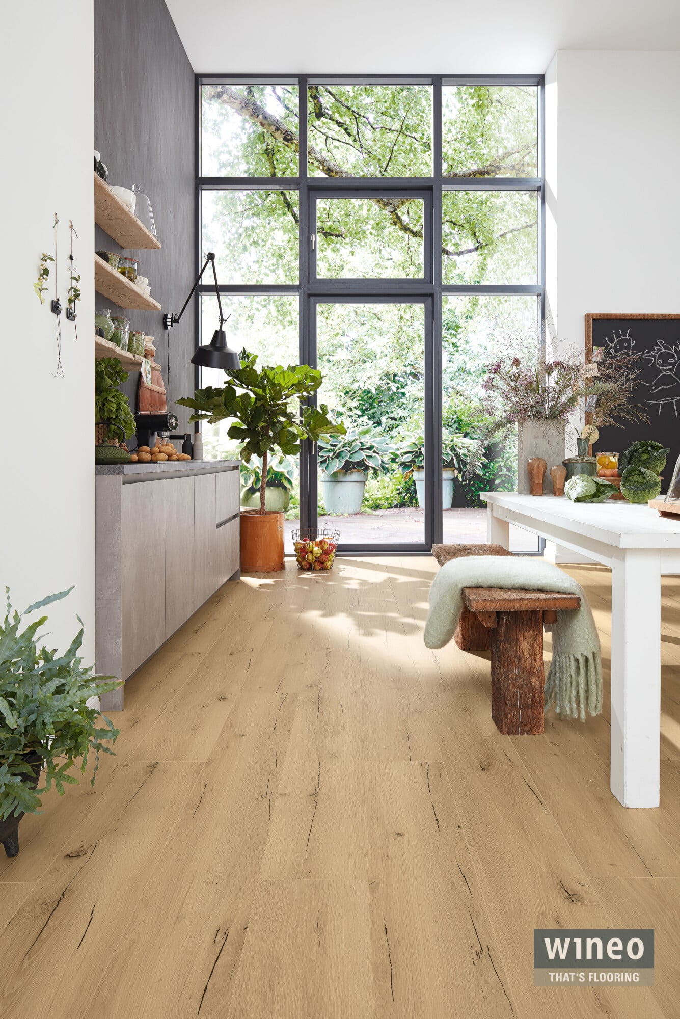 Ein heller, einladender Raum mit großen Fenstern, die viel Tageslicht hereinlassen. Der Boden ist aus hellem Holz, und die Wände sind in neutralen Tönen gehalten. Auf einer langen, weißen Tischfläche liegen frisches Gemüse und Obst, während eine moderne Küchenzeile mit offenen Regalen und einer eleganten Lampe im Hintergrund zu sehen ist. Pflanzen und dekorative Elemente verleihen dem Raum eine lebendige Atmosphäre.