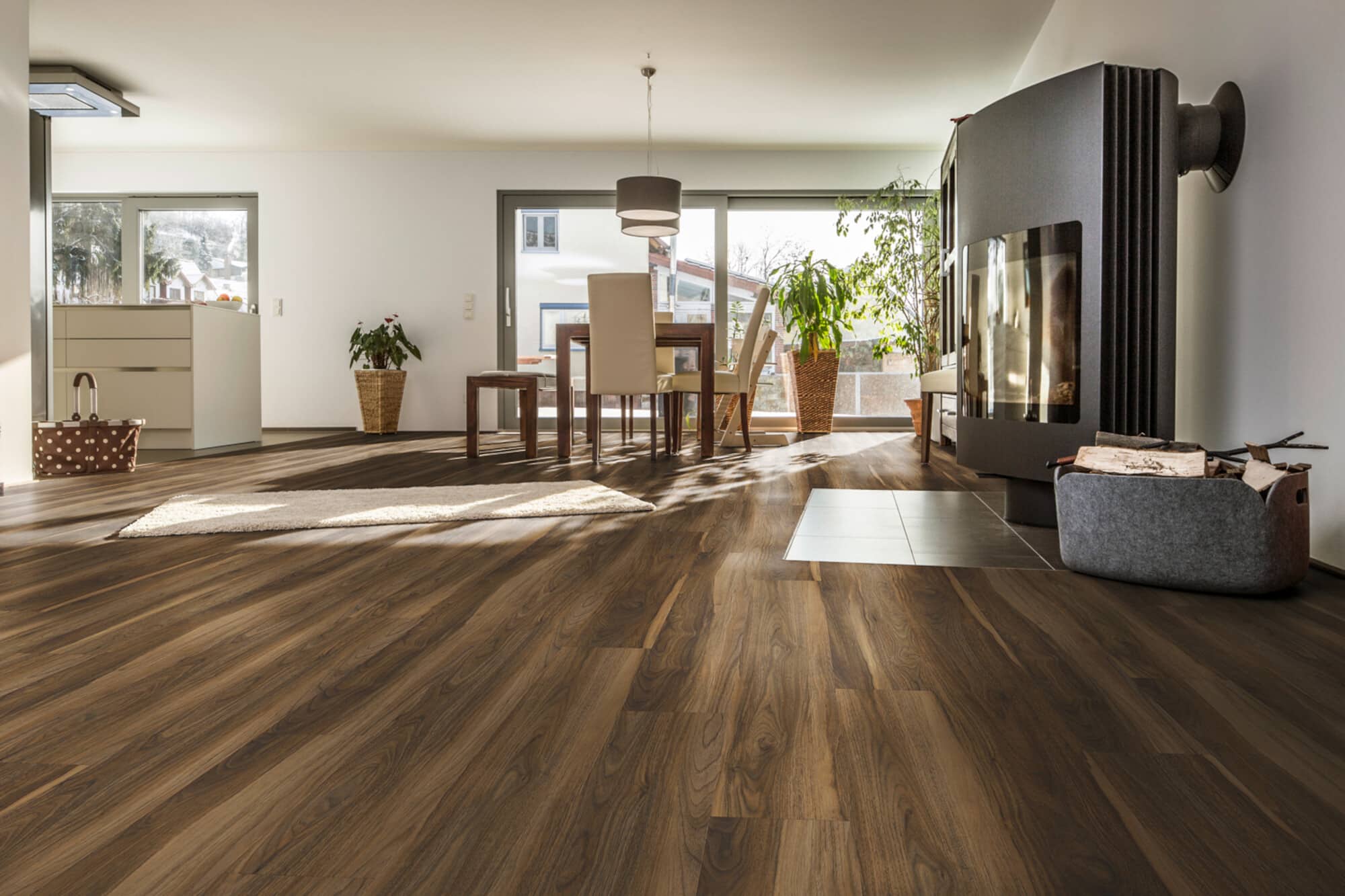 Ein modern eingerichteter Wohnraum mit Holzboden, einem Esstisch mit Stühlen, einem gemütlichen Kamin und großen Fenstern, die viel Tageslicht hereinlassen. Pflanzen und ein Teppich ergänzen die Einrichtung.