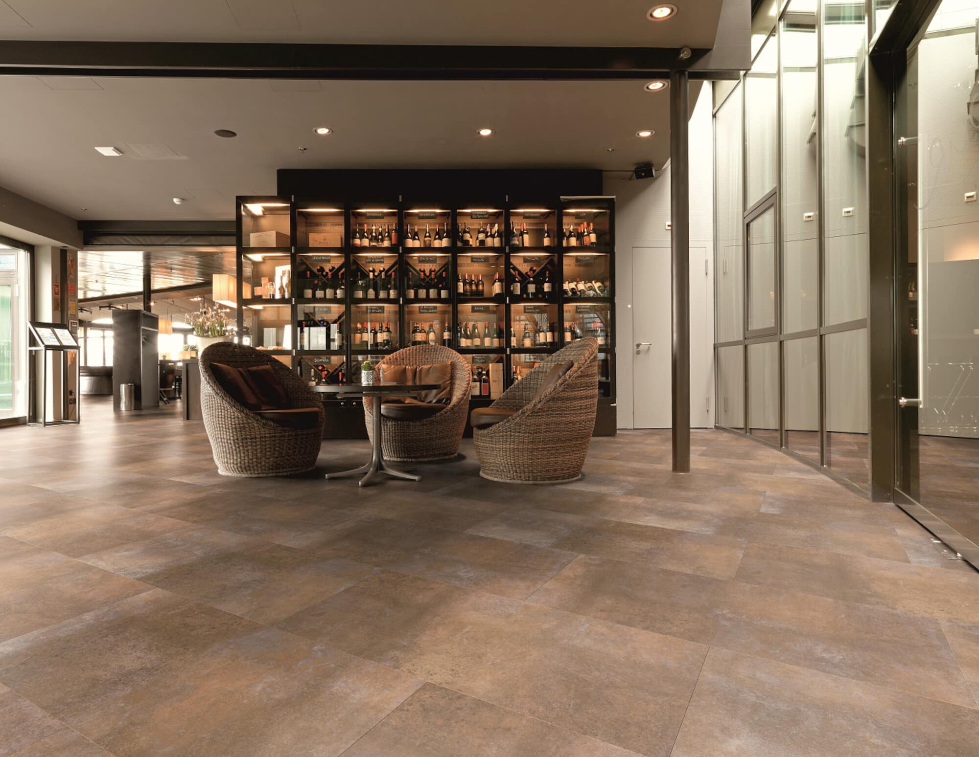 Gemütlicher Lounge-Bereich mit zwei geflochtenen Sesseln und einem kleinen Tisch, umgeben von einer eleganten Bar, die eine Auswahl an Flaschen präsentiert. Der Boden ist in warmen Erdtönen gehalten und das Licht ist sanft und einladend.