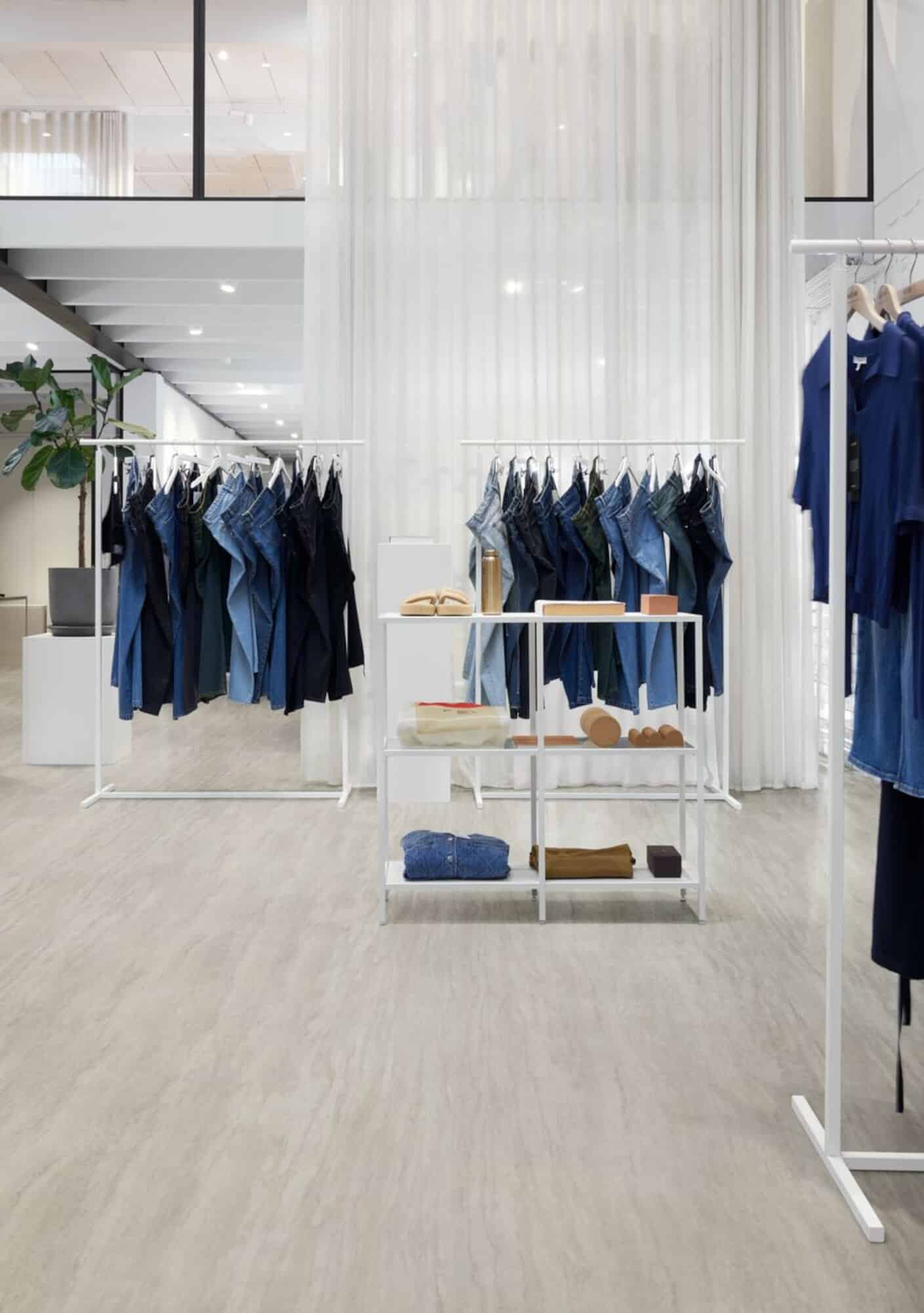 Ein moderner Bekleidungsladen mit weißen Kleiderständern, die verschiedene Jeans und Oberteile in Blau- und Schwarztönen präsentieren. Im Hintergrund sind helle Vorhänge und eine gemütliche Atmosphäre zu sehen. Auf einem Regal sind zusätzlich einige Kleidungsstücke und Accessoires ausgestellt.