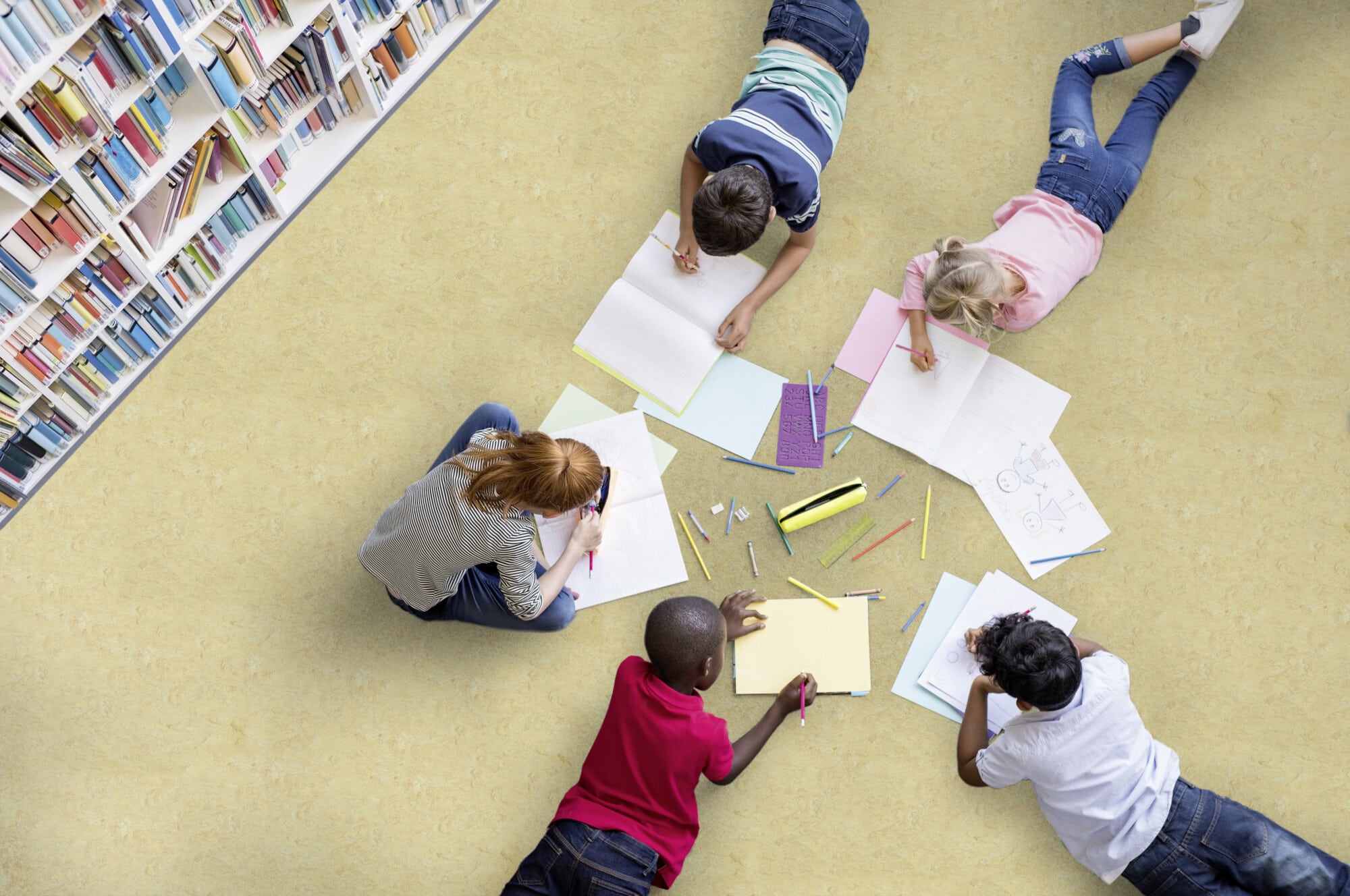 Eine Gruppe von Kindern sitzt auf dem Boden einer Bibliothek und zeichnet in Notizhefte. Um sie herum liegen verschiedene Blätter und Buntstifte. Im Hintergrund sind Regale mit Bücher zu sehen.