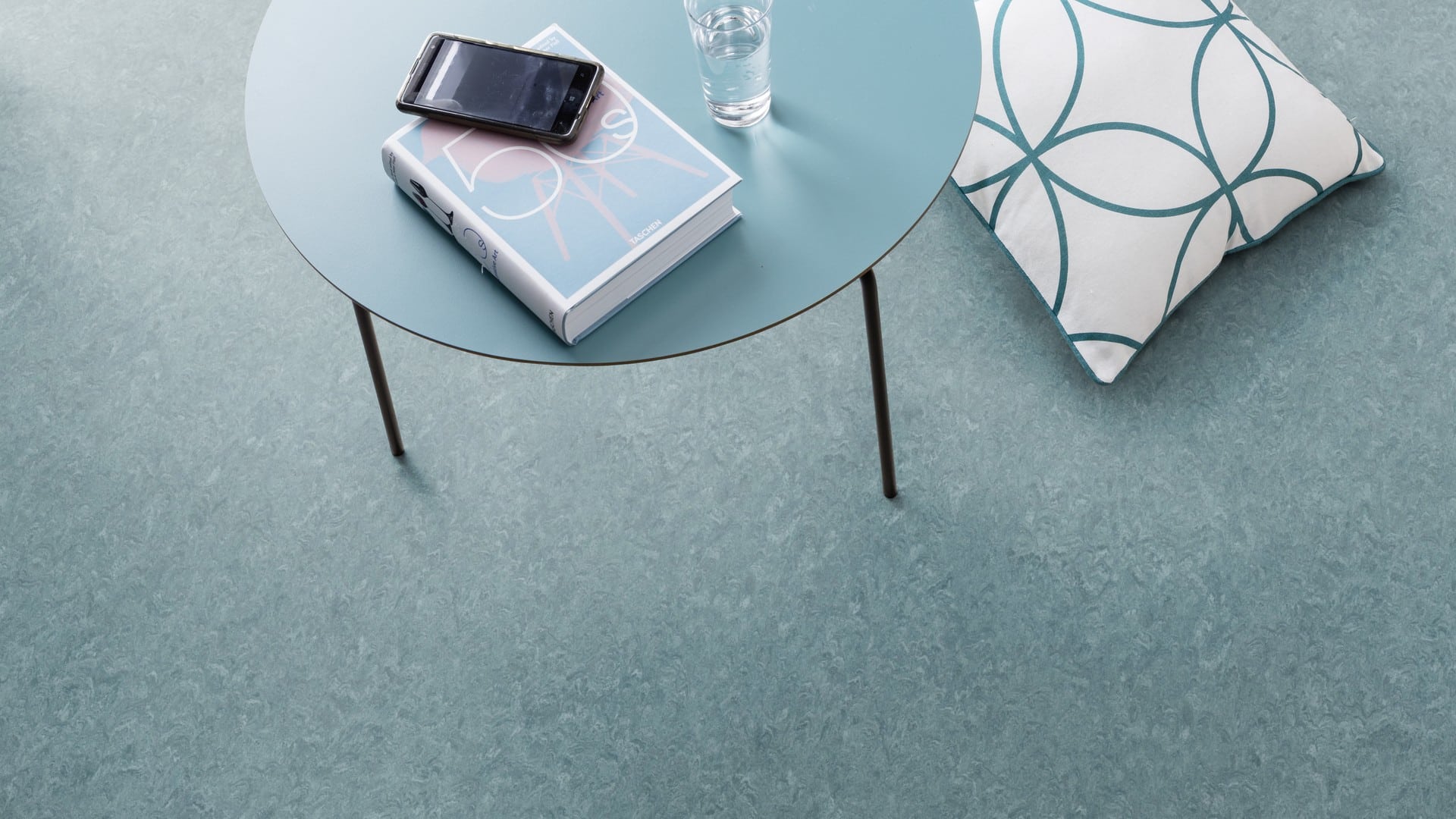 Ein runder, blauer Tisch mit einem Buch, einem Smartphone und einem Glas Wasser darauf, daneben ein Kissen mit einem geometrischen Muster auf einem hellblauen Boden.