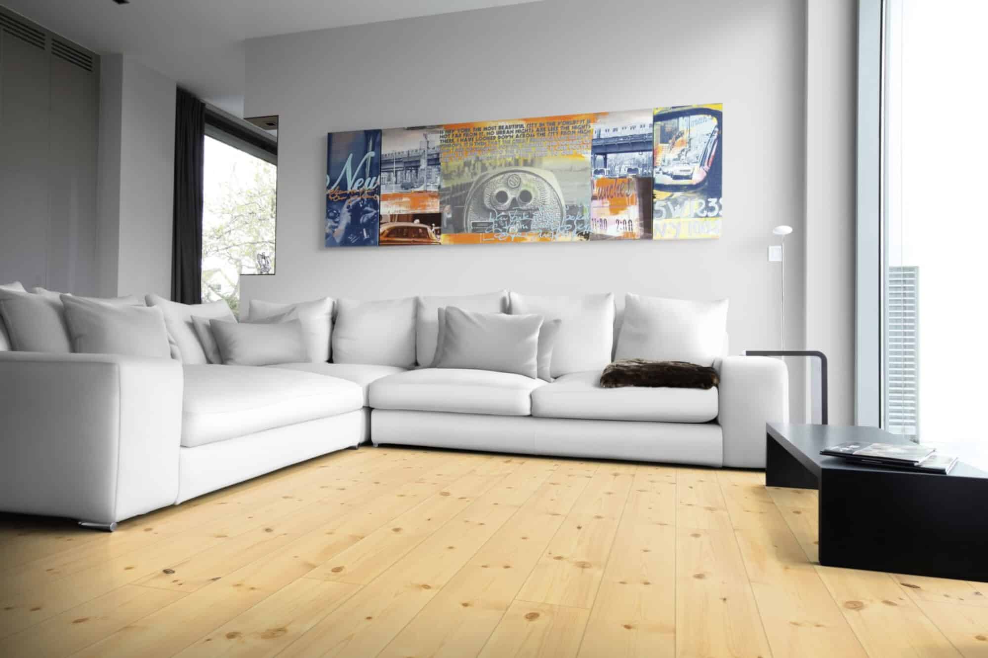 Ein modernes Wohnzimmer mit einem weißen, L-förmigen Sofa, das mit vielen Kissen dekoriert ist. An der Wand hängt ein großformatiges, buntes Kunstwerk, während der Boden aus hellem Holz besteht. Große Fenster lassen viel Tageslicht herein. Ein schwarzer Couchtisch steht im Vordergrund, auf dem einige Zeitschriften liegen.