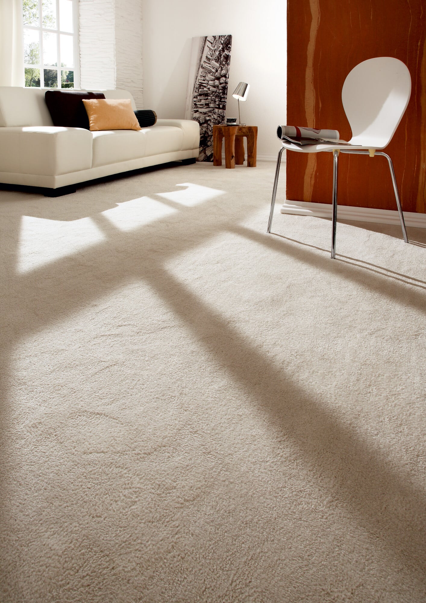 Ein heller, moderner Wohnraum mit einem weißen Sofa, einem Holztisch und einem weißen Stuhl. Der Boden ist mit einem hellen Teppich bedeckt, und Lichtstrahlen fallen durch ein Fenster, das eine warme Atmosphäre schafft. An der Wand befindet sich ein orangefarbener Akzent und ein Bild mit einer Stadtansicht.