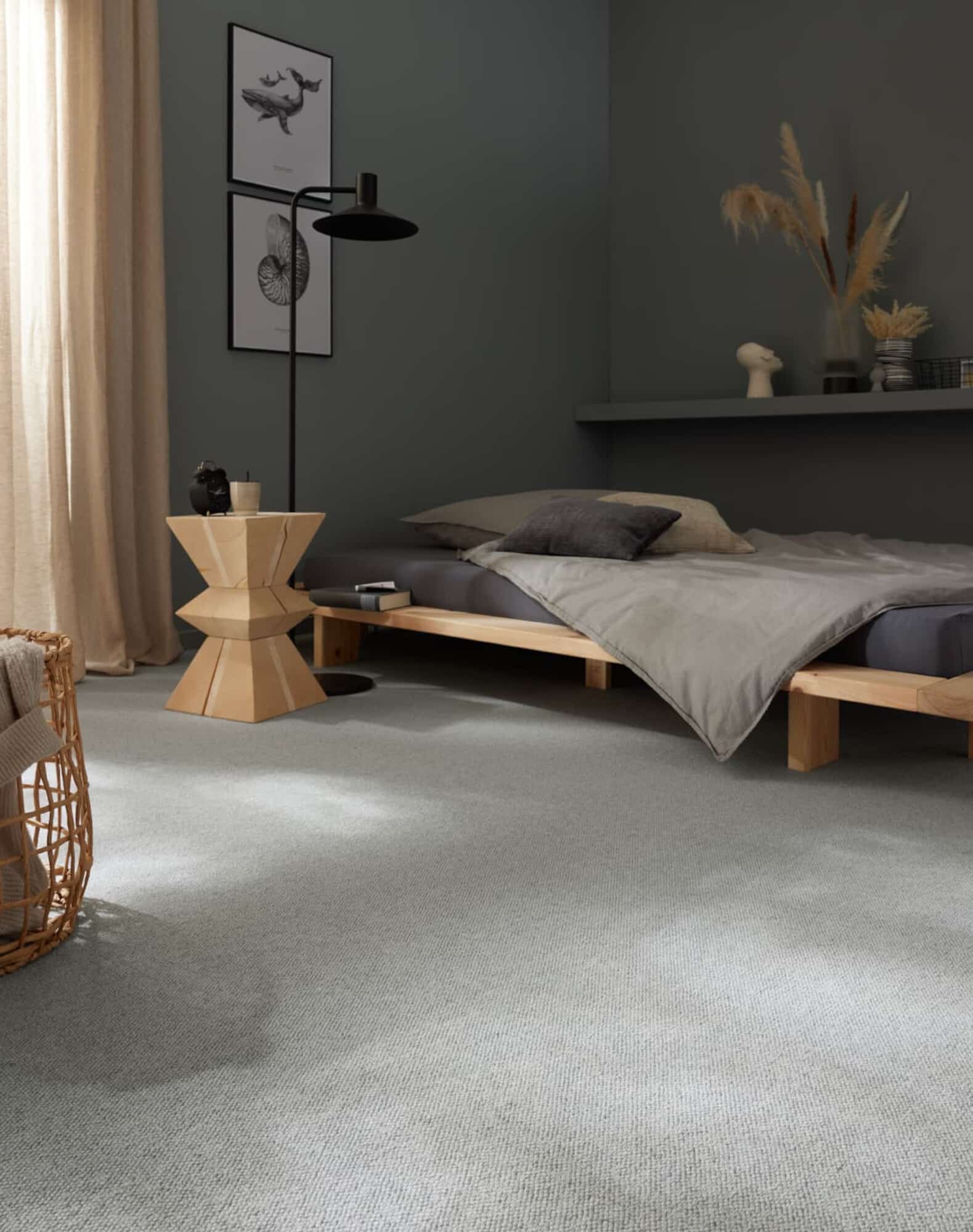Ein modern eingerichtetes Schlafzimmer mit einem niedrigen Bett aus Holz, grauer Bettwäsche und einer Decke. Neben dem Bett steht ein stilvoller Holztisch mit einer schwarzen Lampe und dekorativen Elementen. An der Wand hängen zwei Bilder, und eine Ecke des Raumes ist mit einem großen Korb und einer Pflanze dekoriert. Der Boden ist mit einem hellen Teppich ausgelegt.