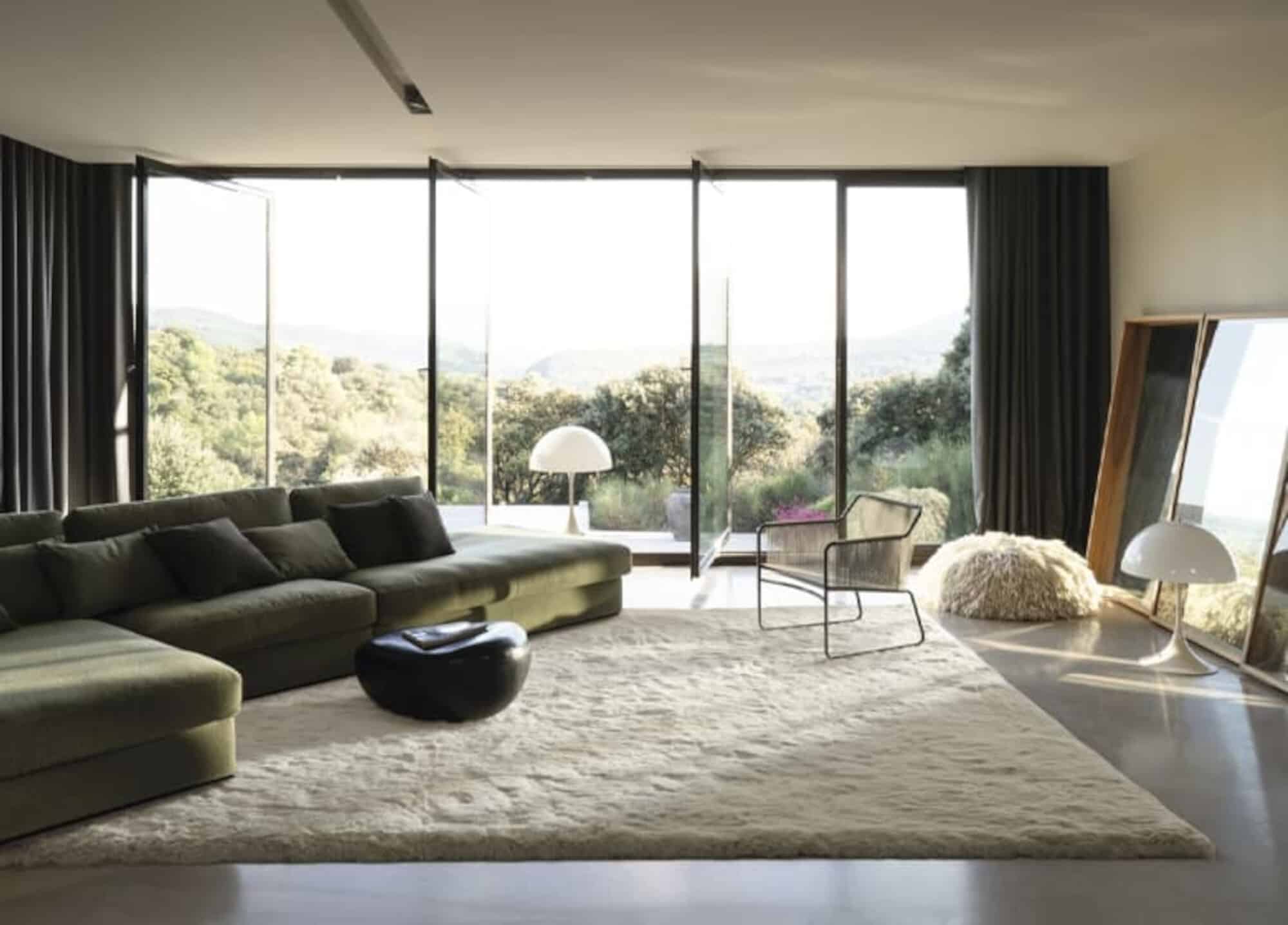 Ein modernes Wohnzimmer mit einer großen, grünen Couch, einem runden schwarzen Tisch und einem Teppich auf einem hellen Boden. Große Fenster bieten einen Blick auf die grüne Landschaft im Freien. Ein elegischer Stuhl und eine Lampe ergänzen die Einrichtung.
