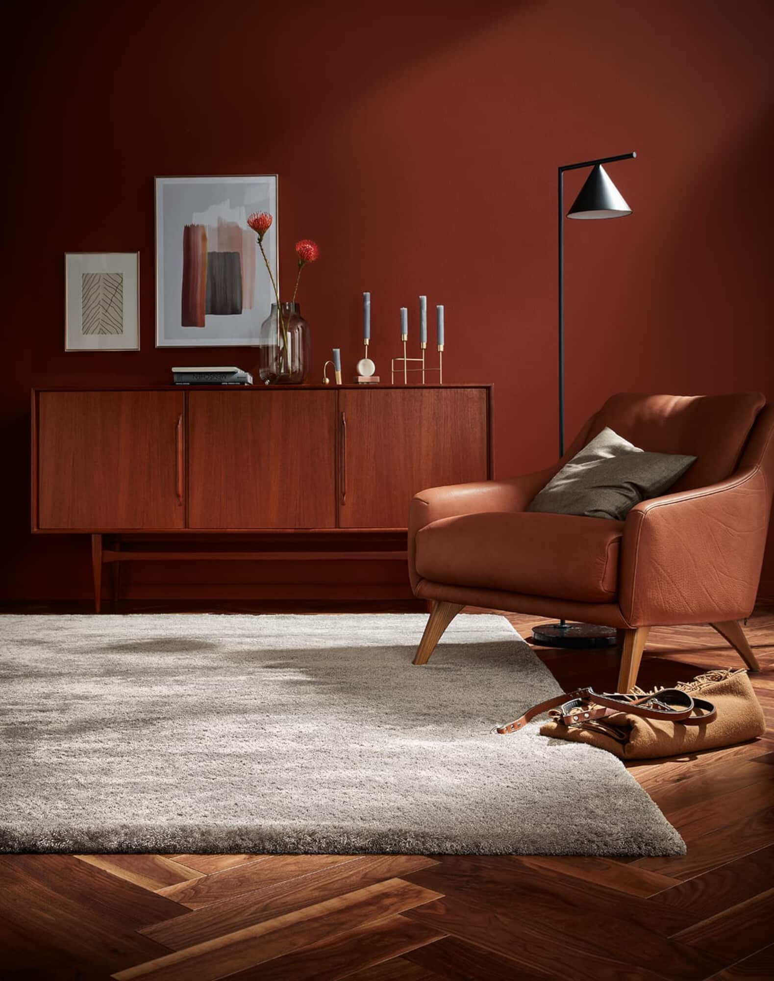 Ein stilvoll eingerichteter Raum mit einer eleganten, braunen Lederarmchair, einem modernen Holzsideboard und einer sanften, grauen Teppichfläche. An der Wand befinden sich Kunstwerke in sanften Farben und eine Stehlampe mit einem minimalistischen Design.