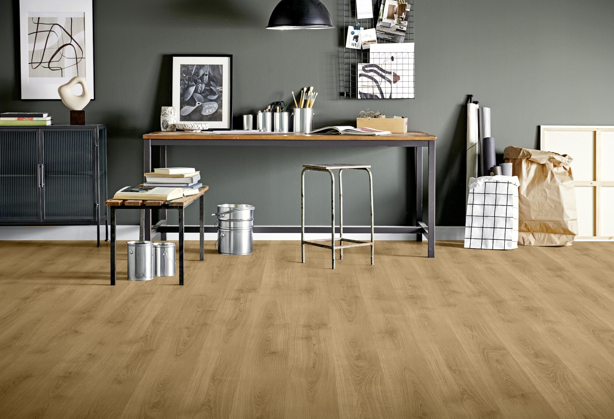 Ein moderner Arbeitsbereich mit einem Holzfußboden, einem langen Tisch aus Holz und Metall, einem Hocker, verschiedenen Büchern und Deko-Elementen. An der Wand hängen Bilder und eine Drahtgittertafel, während einige Materialien in der Nähe aufbewahrt werden.