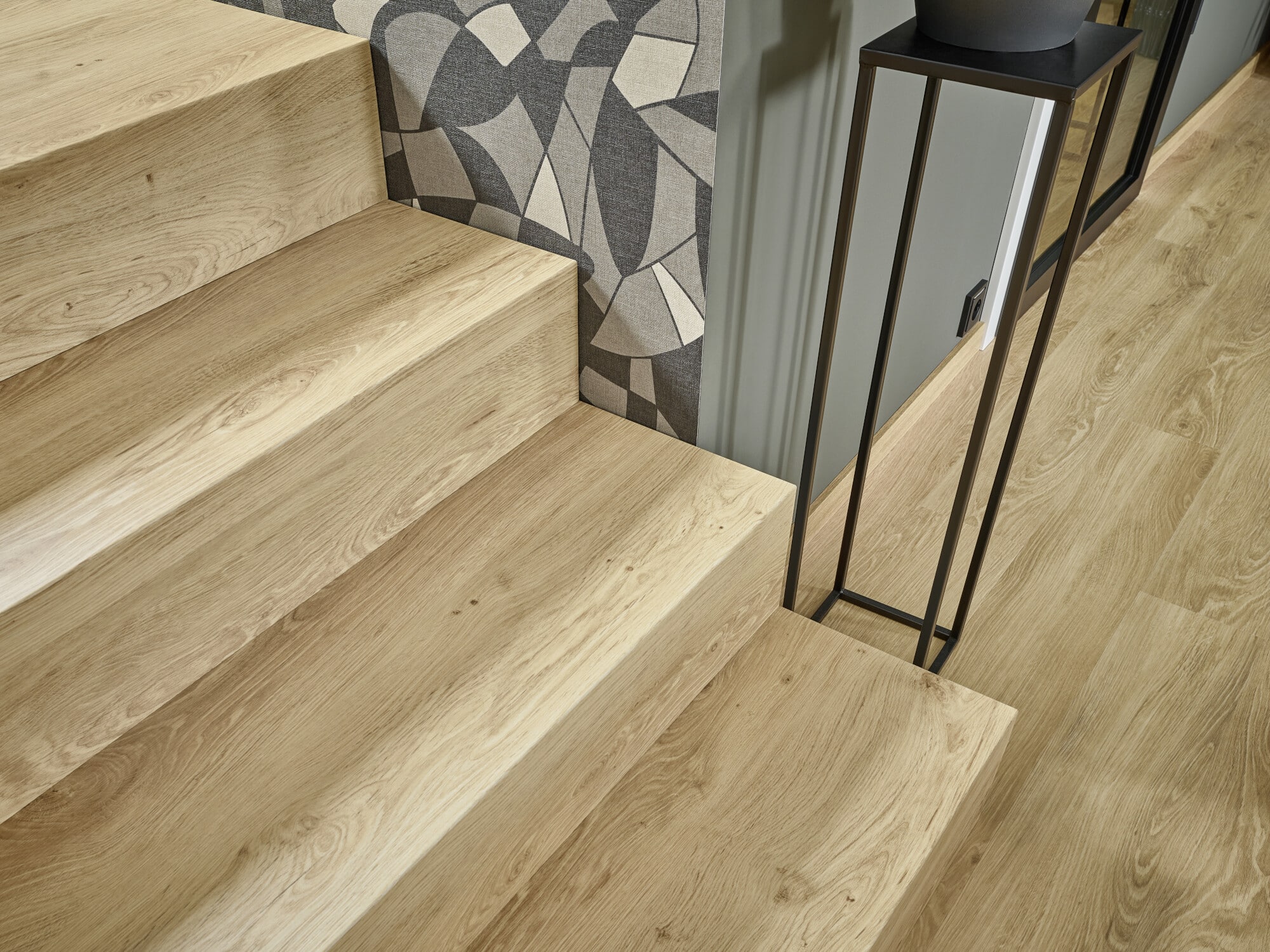 Holztreppe mit hellen Stufen, die zu einem modernen Raum führen. An der Wand ist ein geometrisches Tapetenmuster zu sehen, und neben der Treppe steht ein schlanker, schwarzer Pflanzenständer. Der Boden ist ebenfalls aus hellem Holz.