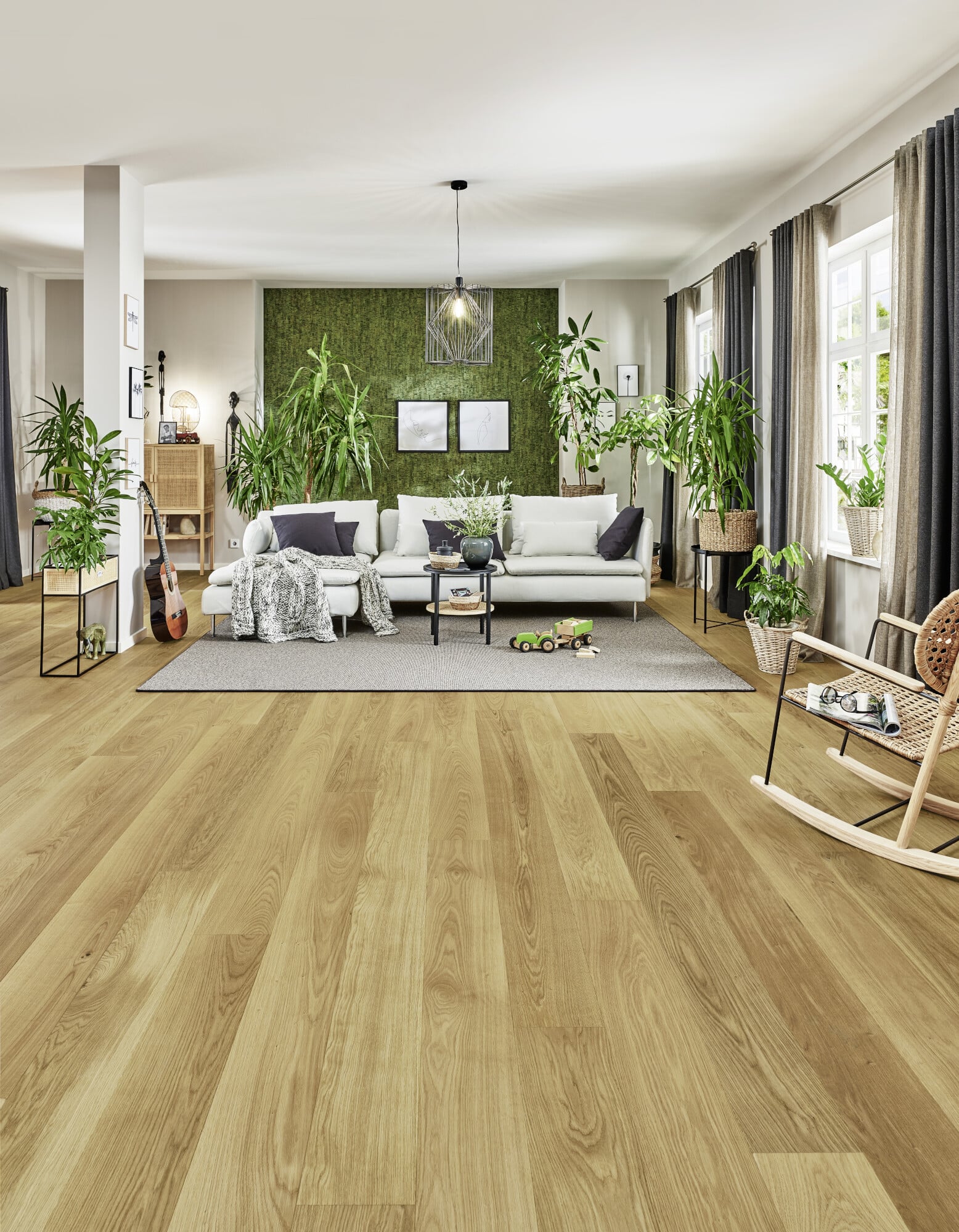 Ein helles, modernes Wohnzimmer mit einem gemütlichen Sofa, einem runden Tisch und vielen Pflanzen. Der Boden ist aus hellem Holz, und an der Wand hängt eine grüne Pflanze. Große Fenster lassen viel Licht herein. Ein Teppich und eine Schaukelstuhl ergänzen die Einrichtung.