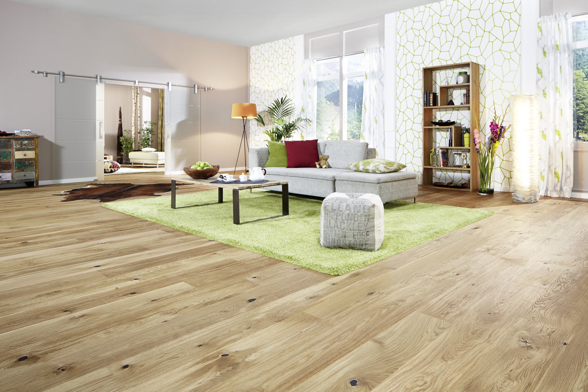 Heller, einladender Wohnraum mit Holzfußboden, einem grauen Sofa, einem Couchtisch und einem grünen Teppich. Große Fenster sorgen für viel Tageslicht, und dekorative Elemente wie Pflanzen und eine Stehlampe verleihen dem Raum eine gemütliche Atmosphäre.