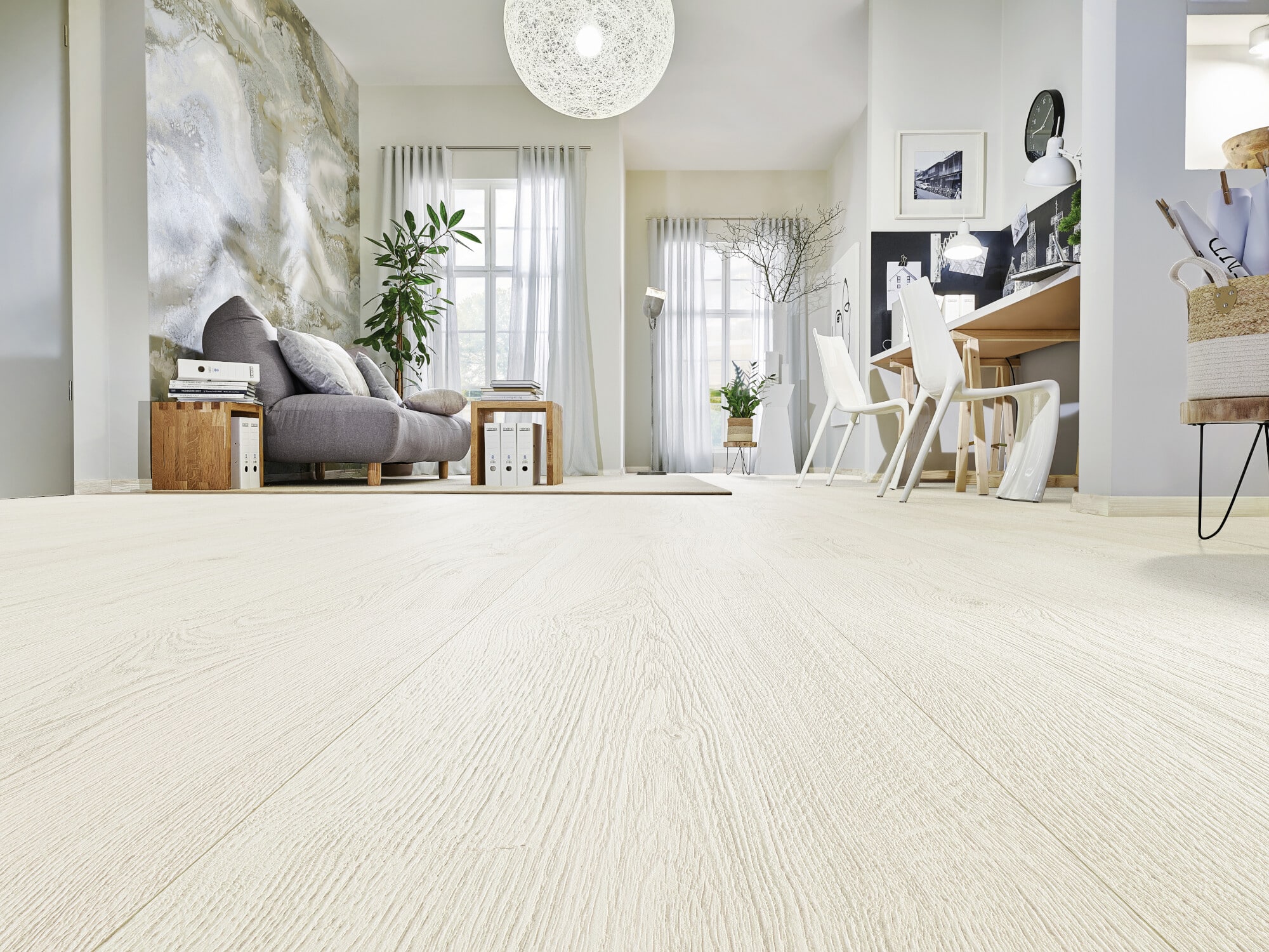 Ein modern eingerichtetes Wohnzimmer mit einem grauen Sofa, Holzmöbeln und einem großen Fenster, das viel Tageslicht hereinlässt. Der Boden ist aus hellem Holz, und im Hintergrund sind ein Esstisch mit weißen Stühlen sowie eine dekorative Wandgestaltung sichtbar.