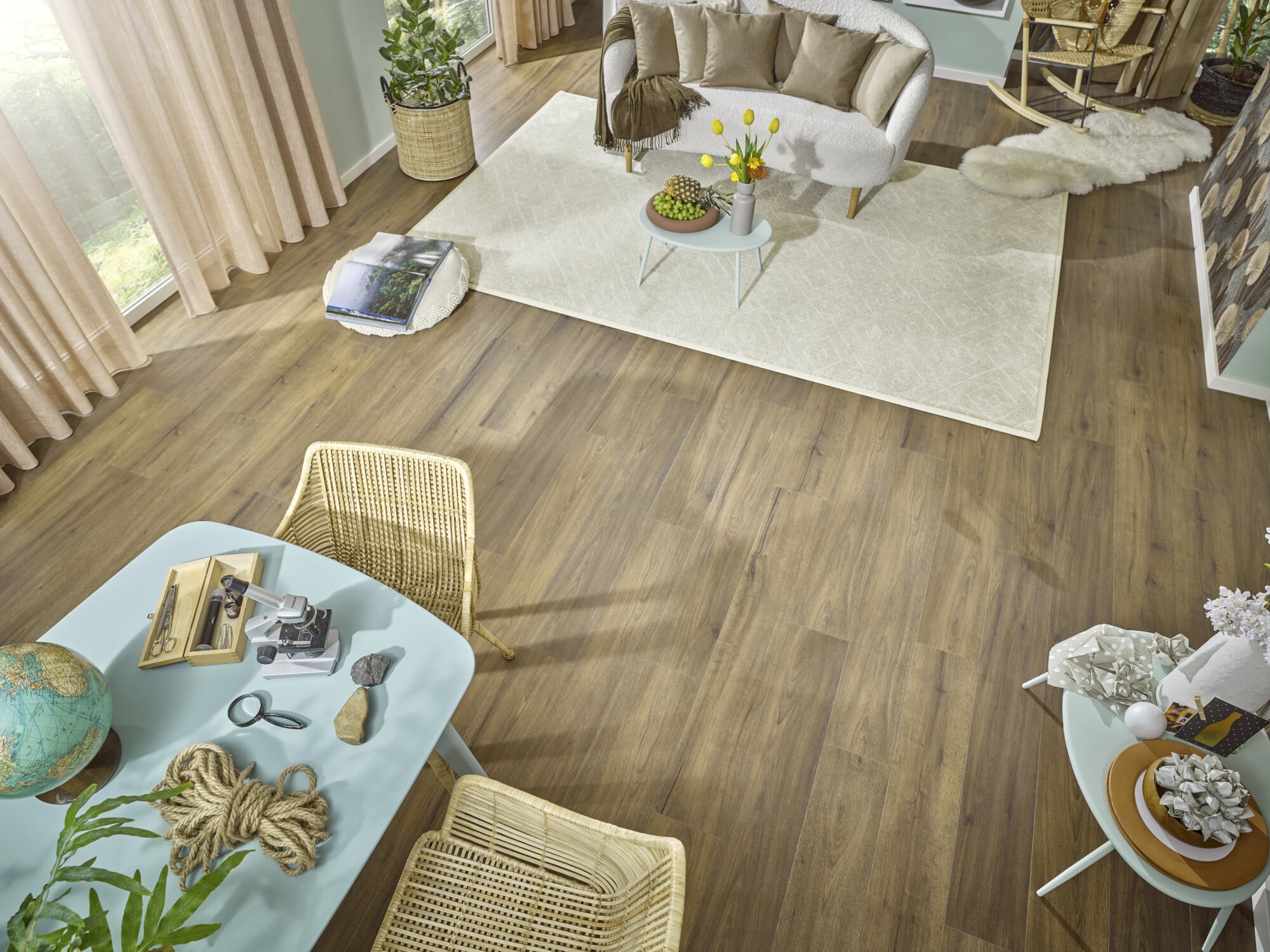 Ein helles und einladendes Wohnzimmer mit einem runden Tisch, zwei Stühlen und einem gemütlichen Sofa. Auf dem Tisch liegen eine Weltkugel, eine Lupe, eine Schere und ein Stein. Der Boden ist aus Holz und es gibt einen weichen Teppich sowie Pflanzen und Dekorationselemente im Raum. Licht strömt durch große Fenster mit Vorhängen.