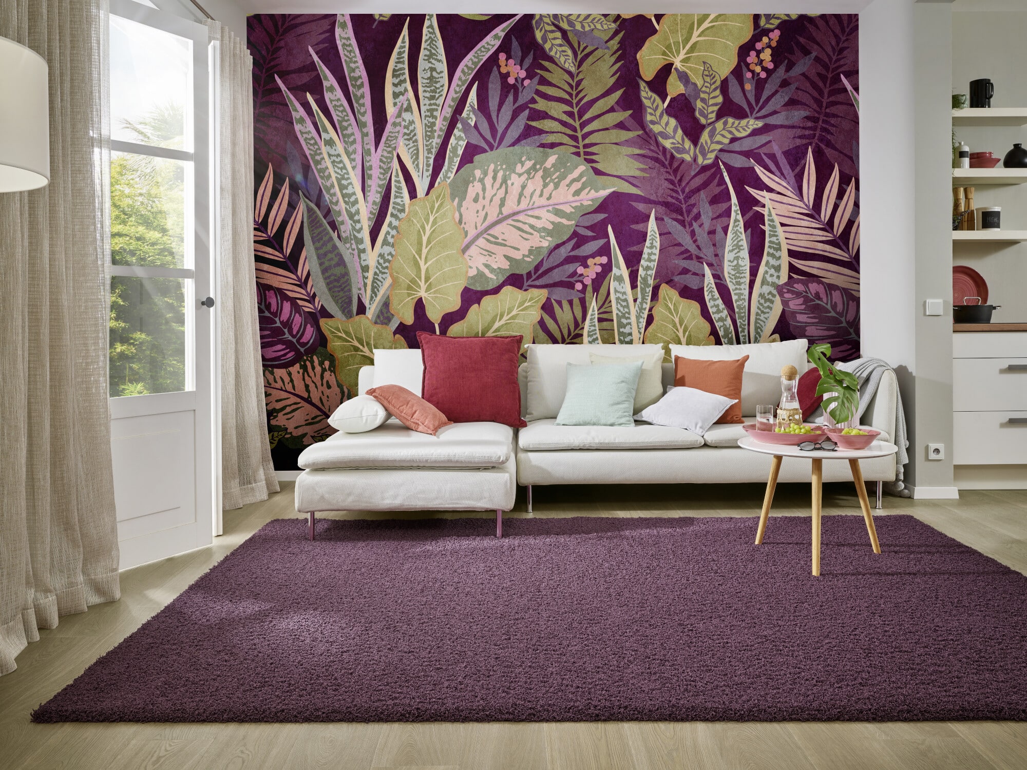Ein modernes Wohnzimmer mit einem weißen Sofa, bunten Kissen und einem runden Tisch. Der Hintergrund ist mit einem großflächigen, bunten Pflanzenmuster dekoriert. Auf dem Boden liegt ein lila Teppich, und ein Fenster lässt Tageslicht herein.