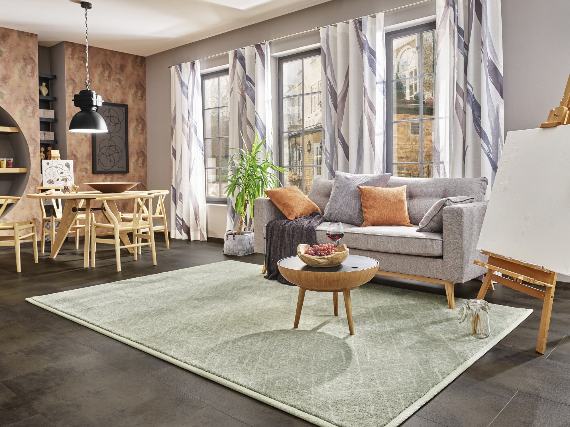 Ein modern eingerichtetes Wohnzimmer mit einem grauen Sofa, bunten Kissen und einem runden Couchtisch. Auf dem Tisch steht eine Schale mit Obst und ein Glas Wein. Der Raum hat große Fenster mit Vorhängen, einen Esstisch aus Holz und einen kleinen grünen Pflanzen. Der Boden ist dunkel und der Teppich ist hellgrün mit einem dezenten Muster. An der Wand steht eine Staffelei.