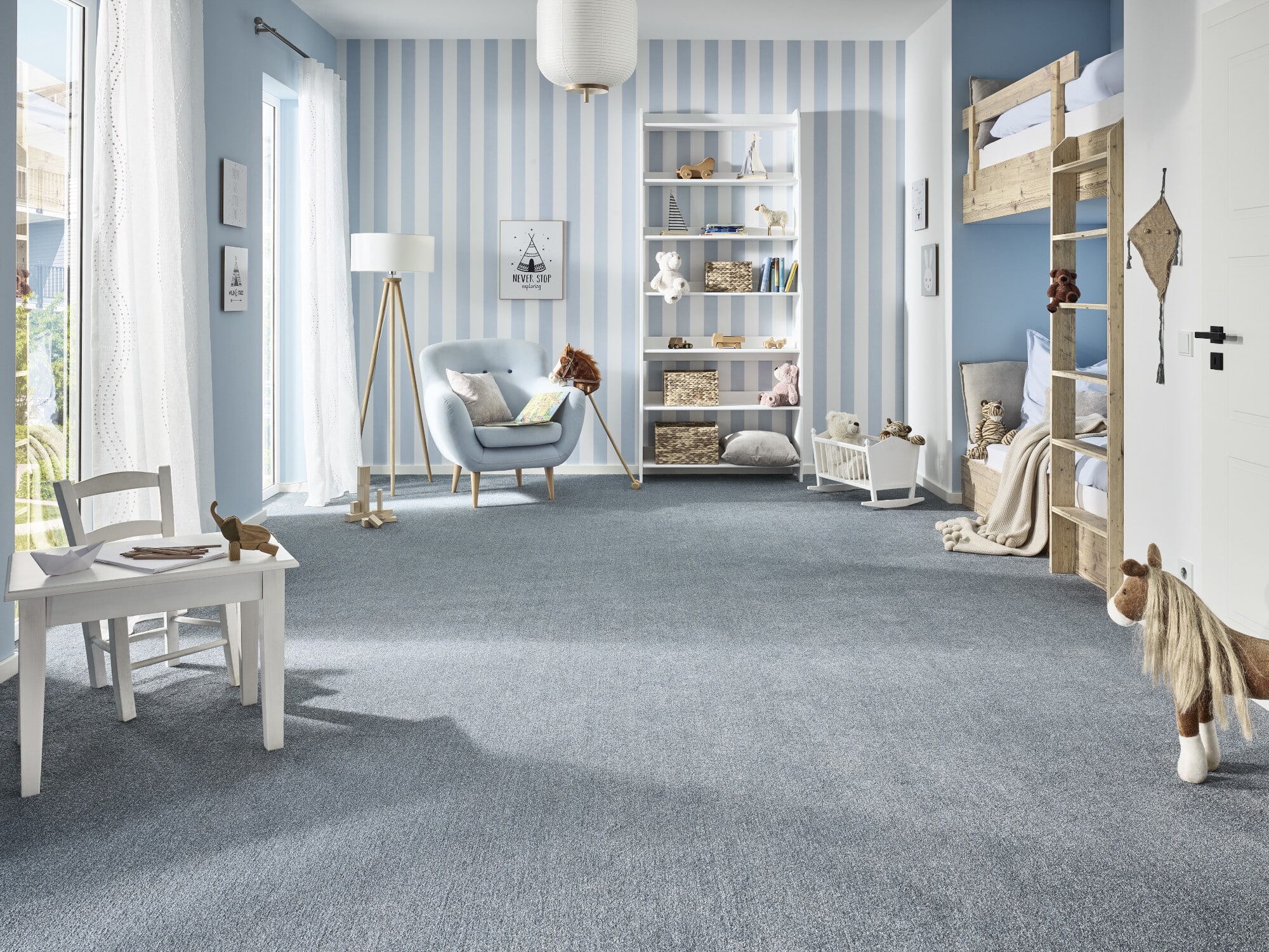 Ein hellblauer Kinderraum mit gestreiften Wänden, einem gemütlichen Sessel, einem weißen Tisch und Stühlen. Im Hintergrund steht ein Hochbett aus Holz mit Kuscheltieren. Ein Teppich in Blau bedeckt den Boden, und große Fenster lassen viel Licht herein.