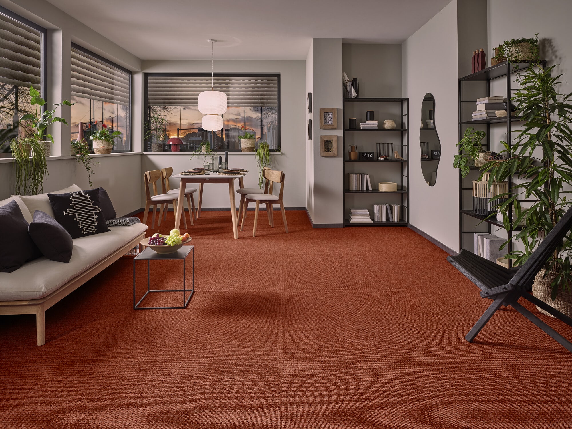 Ein modernes Wohnzimmer mit einem roten Teppich, einer hellen Couch mit Kissen, einem Esstisch mit Stühlen, und großen Fenstern, die den Sonnenuntergang zeigen. Pflanzen schmücken den Raum, und ein offenes Regal mit Dekorationsgegenständen ist ebenfalls sichtbar.