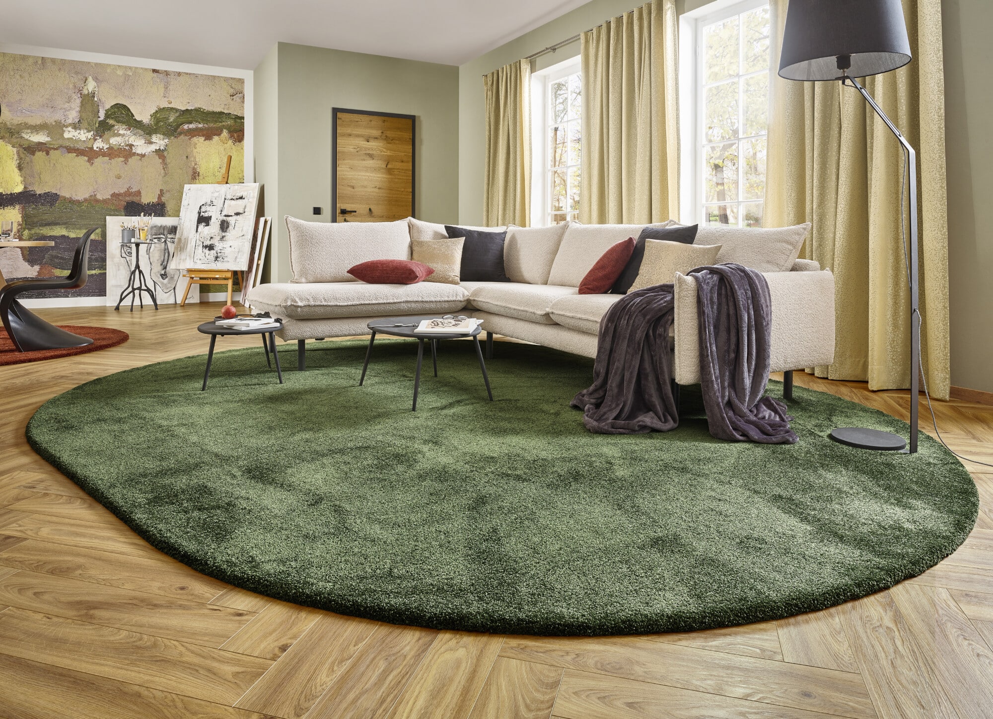 Ein modernes Wohnzimmer mit einem hellen, bequemen Sofa, bunten Kissen und einer gemütlichen Decke. Der Boden ist mit einem runden, grünen Teppich bedeckt, während ein schwarzer Beistelltisch und eine stylische Stehlampe das Ambiente ergänzen. Helle Vorhänge und ein kunstvolles Wandbild verleihen dem Raum eine einladende Atmosphäre.