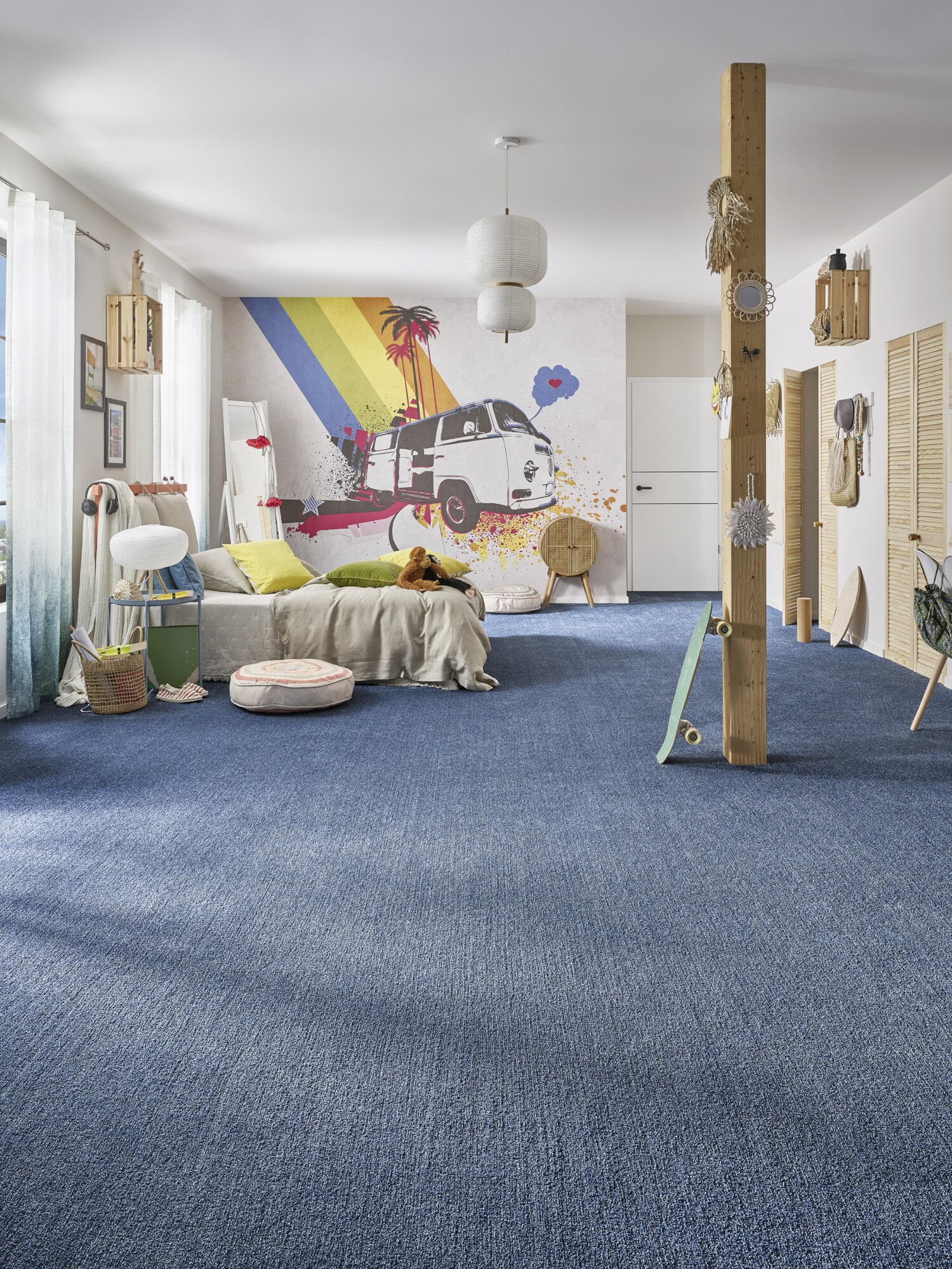 Ein modernes Wohnzimmer mit blauen Teppichboden, einem gemütlichen Bett mit bunten Kissen und einer Wand mit einem farbenfrohen Wandgemälde eines VW-Busses und Palmen. Im Raum befinden sich verschiedene dekorative Elemente wie Pflanzen, eine Stehlampe und ein Holzpfeiler.