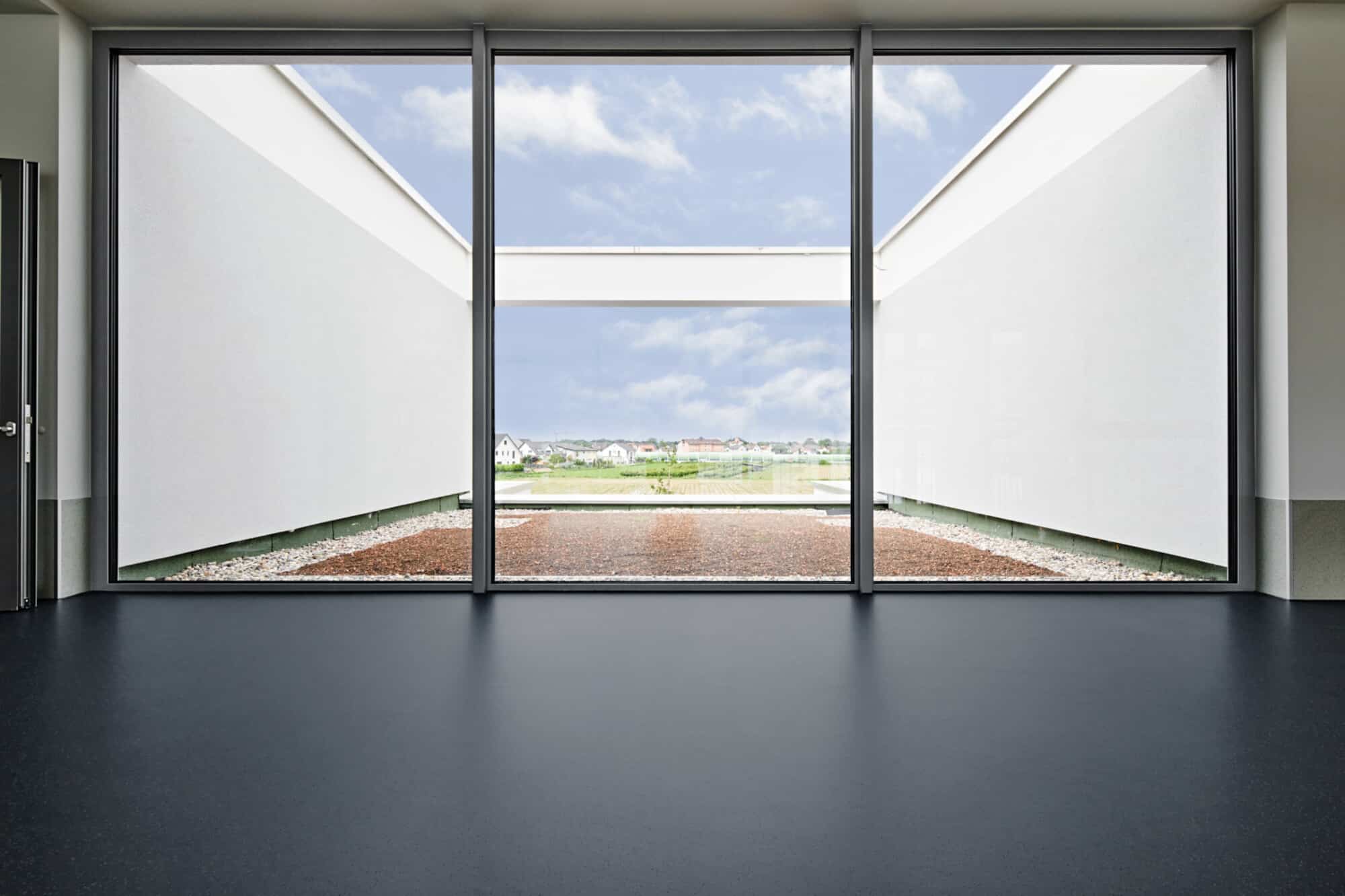 Ein moderner Raum mit großen Fenstern, die einen Blick auf einen begrünten Außenbereich und einen klaren Himmel bieten. Der Boden ist dunkel und glatt, während die Wände weiß sind, was eine helle und luftige Atmosphäre schafft.