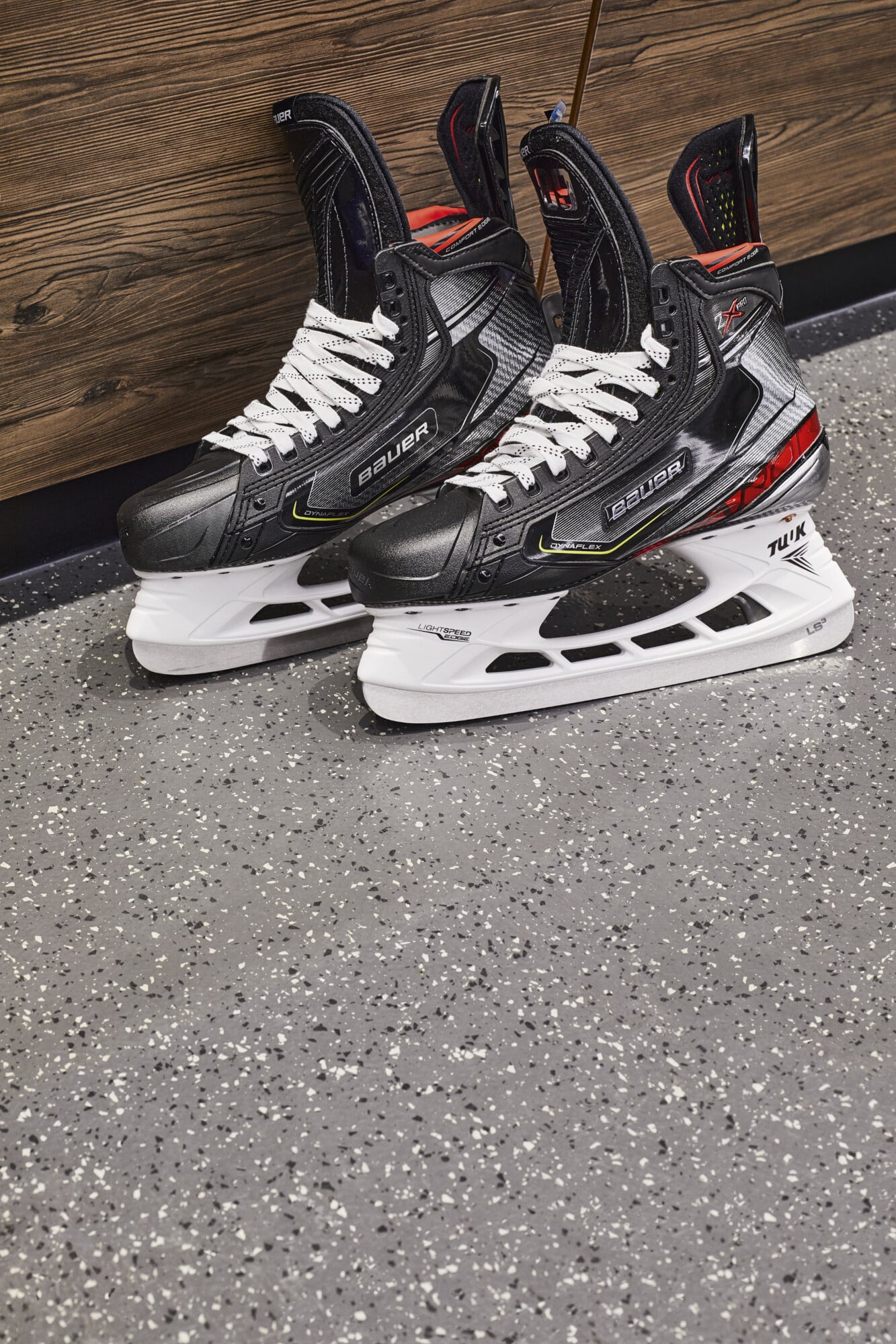 Schwarze Eishockeyschlittschuhe mit weißen Schnürsenkeln stehen auf einem grauen, strukturierten Boden. Im Hintergrund ist eine Holzpaneelwand zu sehen.