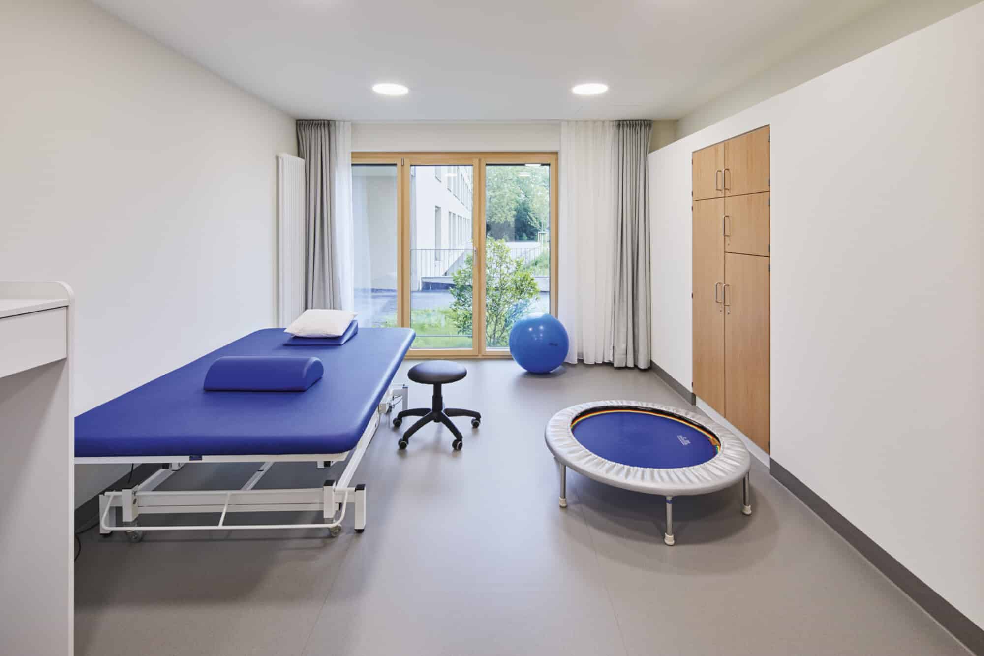 Ein heller Therapieraum mit einem blauen Behandlungstisch, einem Hocker, einem Trampolin und einem großen Fitnessball. Große Fenster lassen Tageslicht herein und bieten einen Blick ins Freie. An der Wand befinden sich Holzschränke für die Aufbewahrung von Materialien.