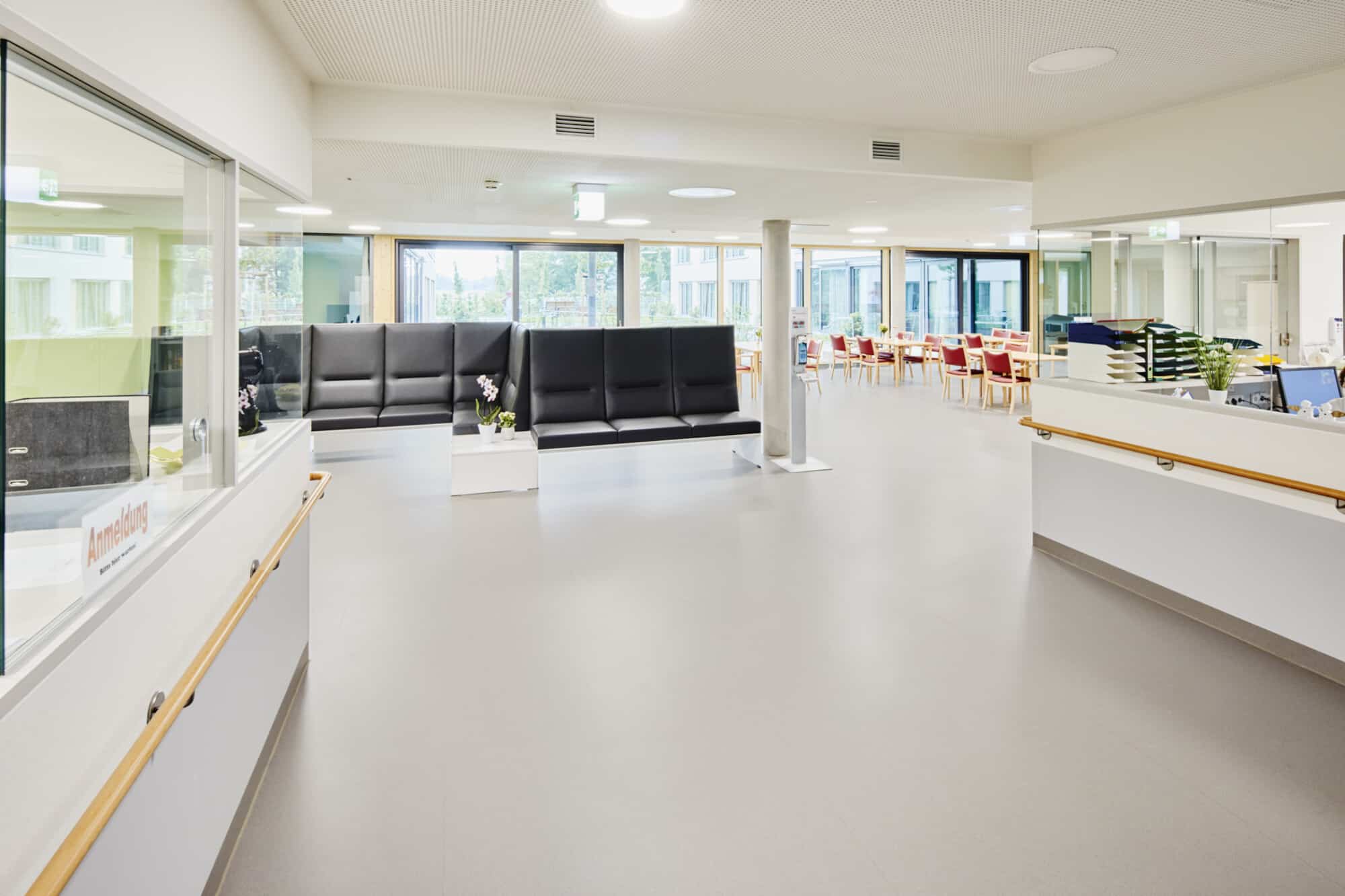 Helle, moderne Empfangshalle mit Sitzgelegenheiten und einem Empfangstresen. Große Fenster sorgen für viel Tageslicht, und im Hintergrund sind Tische mit Stühlen zu sehen.