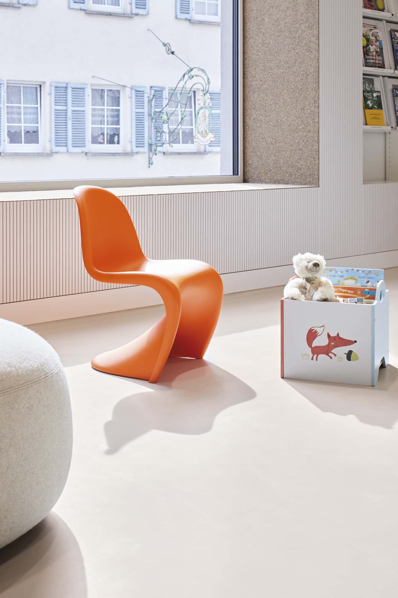 Ein orangefarbener Designstuhl steht neben einer Spielkiste mit einem roten Fuchs-Motiv und einem Stofftier auf einem hellen Boden in einem Raum mit großen Fenstern.