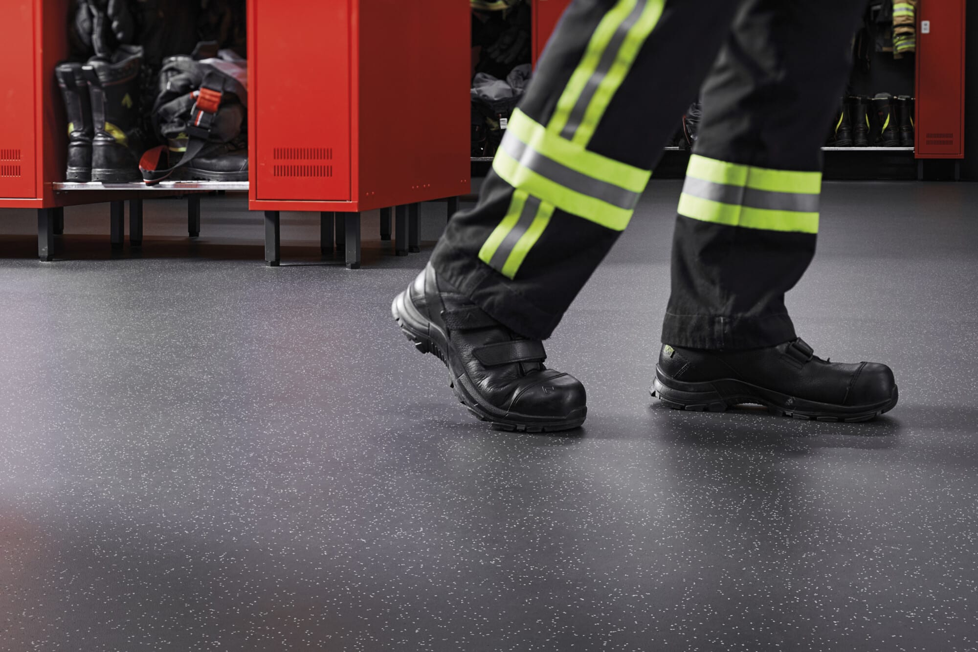 Ein Fuß in schwarzer Arbeitssicherheitsschuhe mit reflektierenden Streifen auf der Hose, der über einen modernen, dunklen Boden in einem Umkleideraum mit roten Spinden geht.