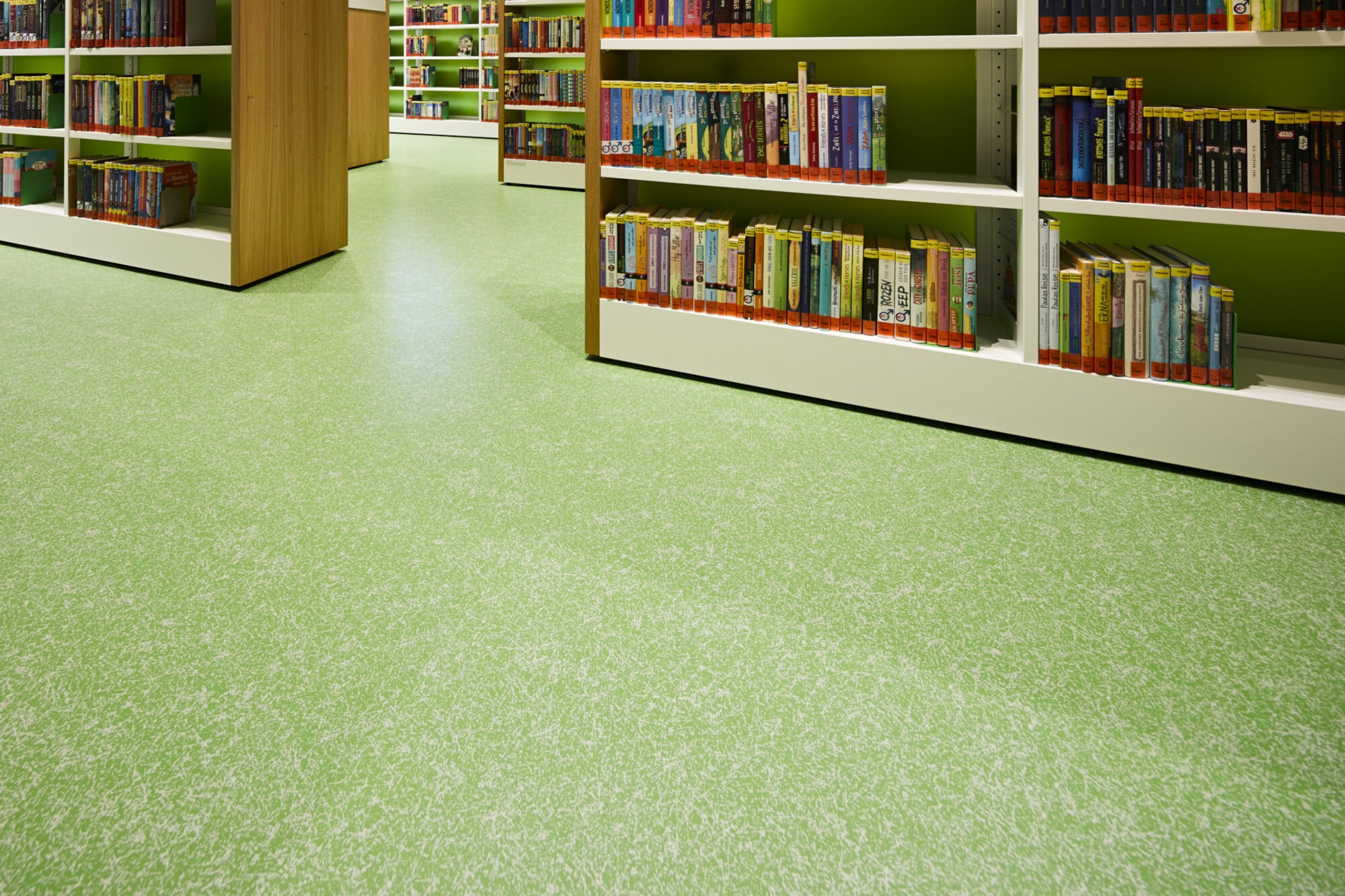 Eine Ansicht des Fußbodens in einer Bibliothek mit grünem, strukturiertem Bodenbelag und Regalen voller Bücher in bunten Umschlägen.