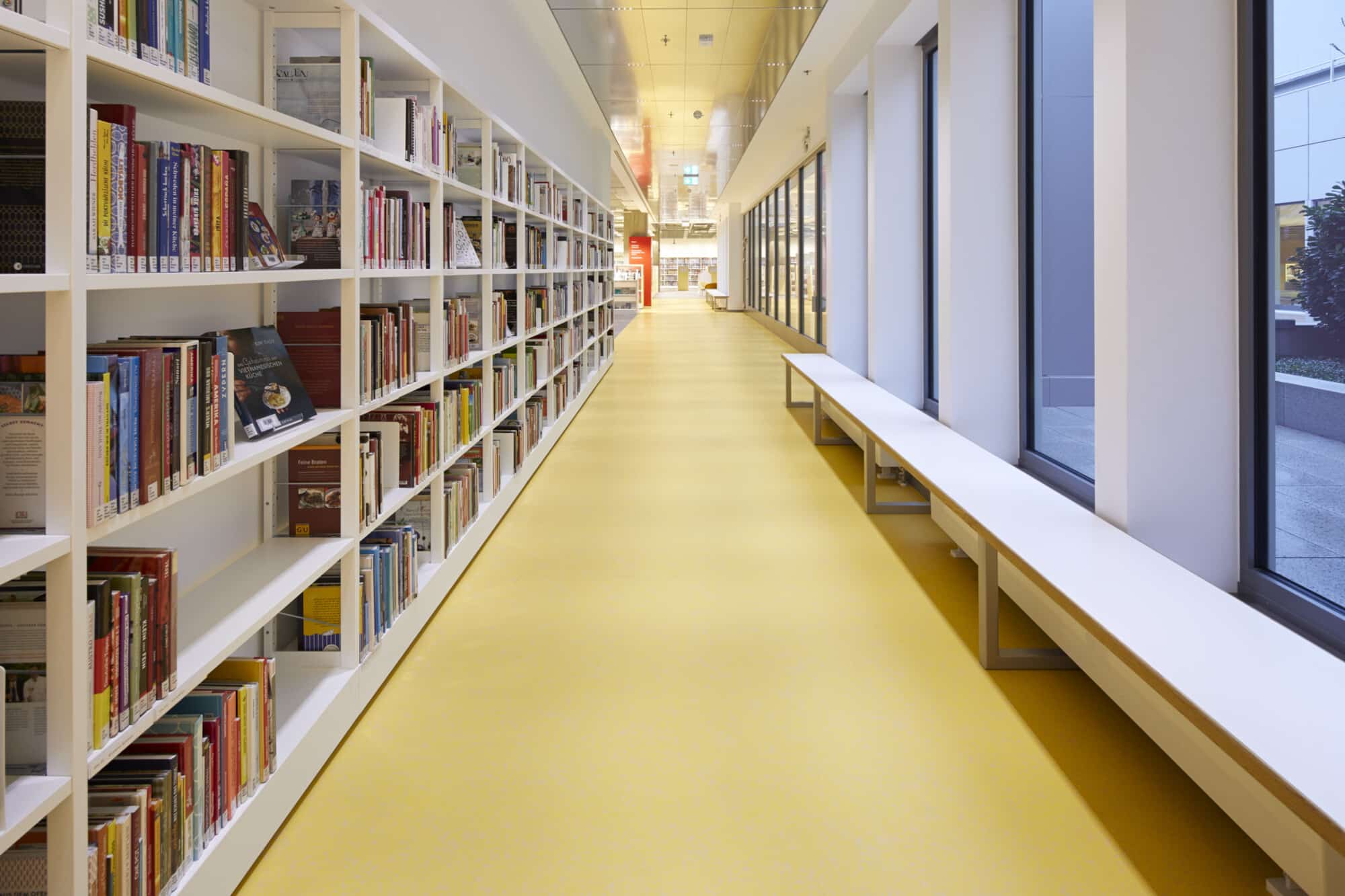 Ein heller, moderner Bibliotheksgang mit weißen Bücherregalen, die mit bunten Büchern gefüllt sind. Der Boden ist in einem warmen Gelbton gehalten, und an der Seite stehen lange, schlichte Sitzbänke. Große Fenster lassen viel Tageslicht herein und bieten einen Blick nach draußen.