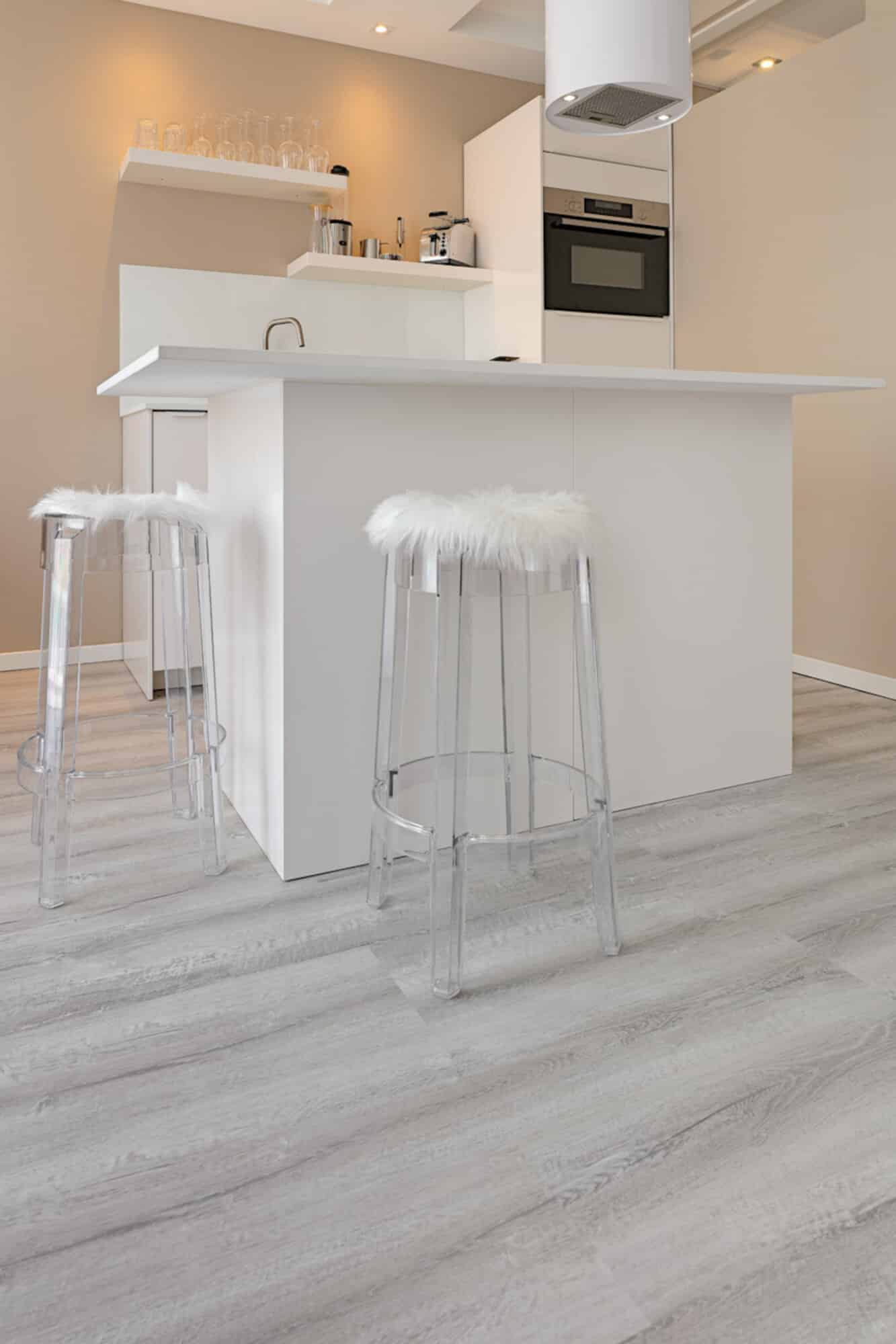 Moderne Küche mit einer weißen Insel, klaren Barhockern mit weißen Fellen und einem hellen, holzähnlichen Boden. Im Hintergrund sind Küchengeräte und Gläser auf einem Regal sichtbar.