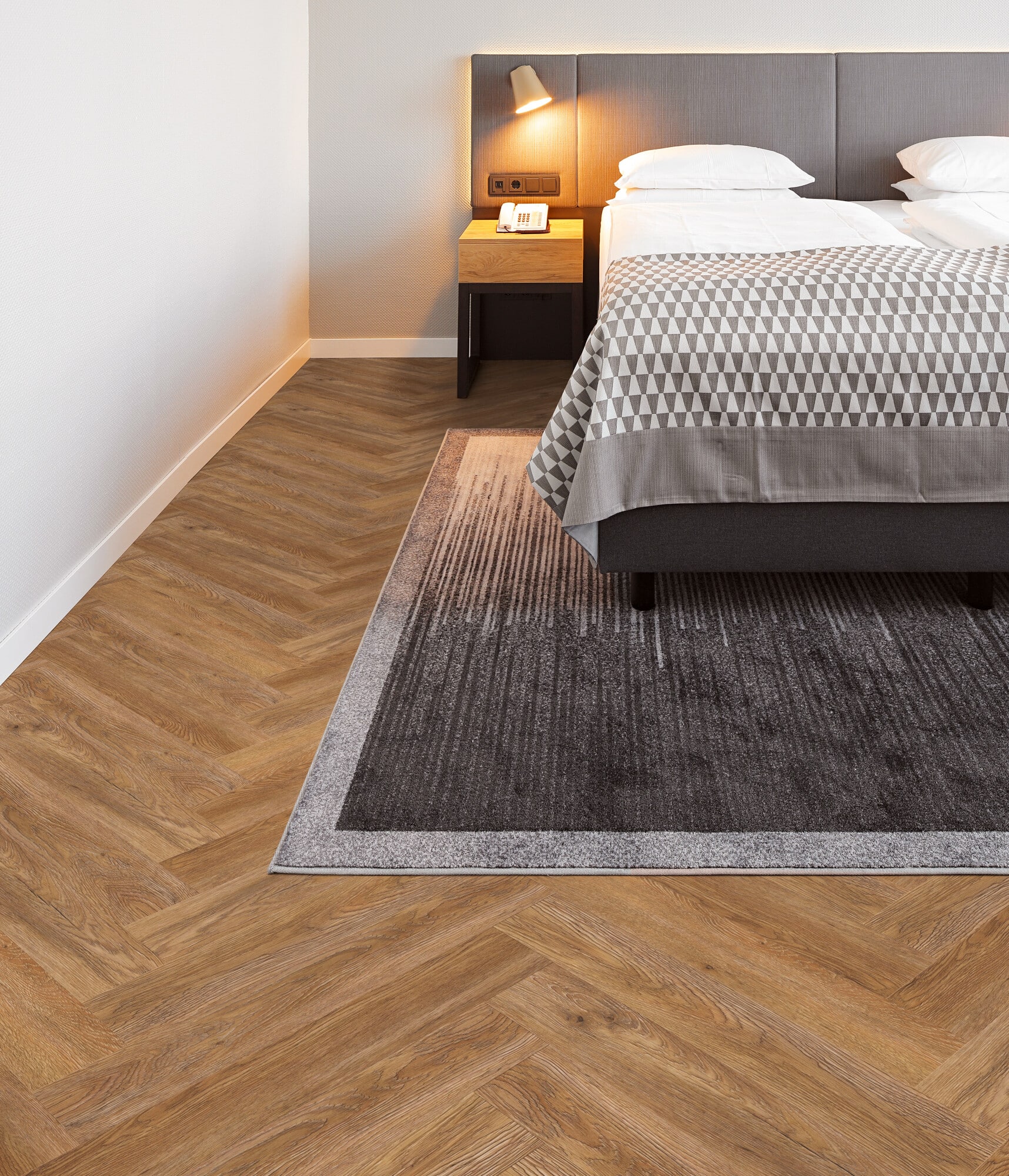 Ein modernes Schlafzimmer mit einem Doppelbett, einem Nachttisch und einer Leselampe. Der Boden ist mit einem hölzernen Fischgrätmuster ausgelegt, während ein grauer Teppich mit einem minimalistischen Design auf dem Boden liegt.