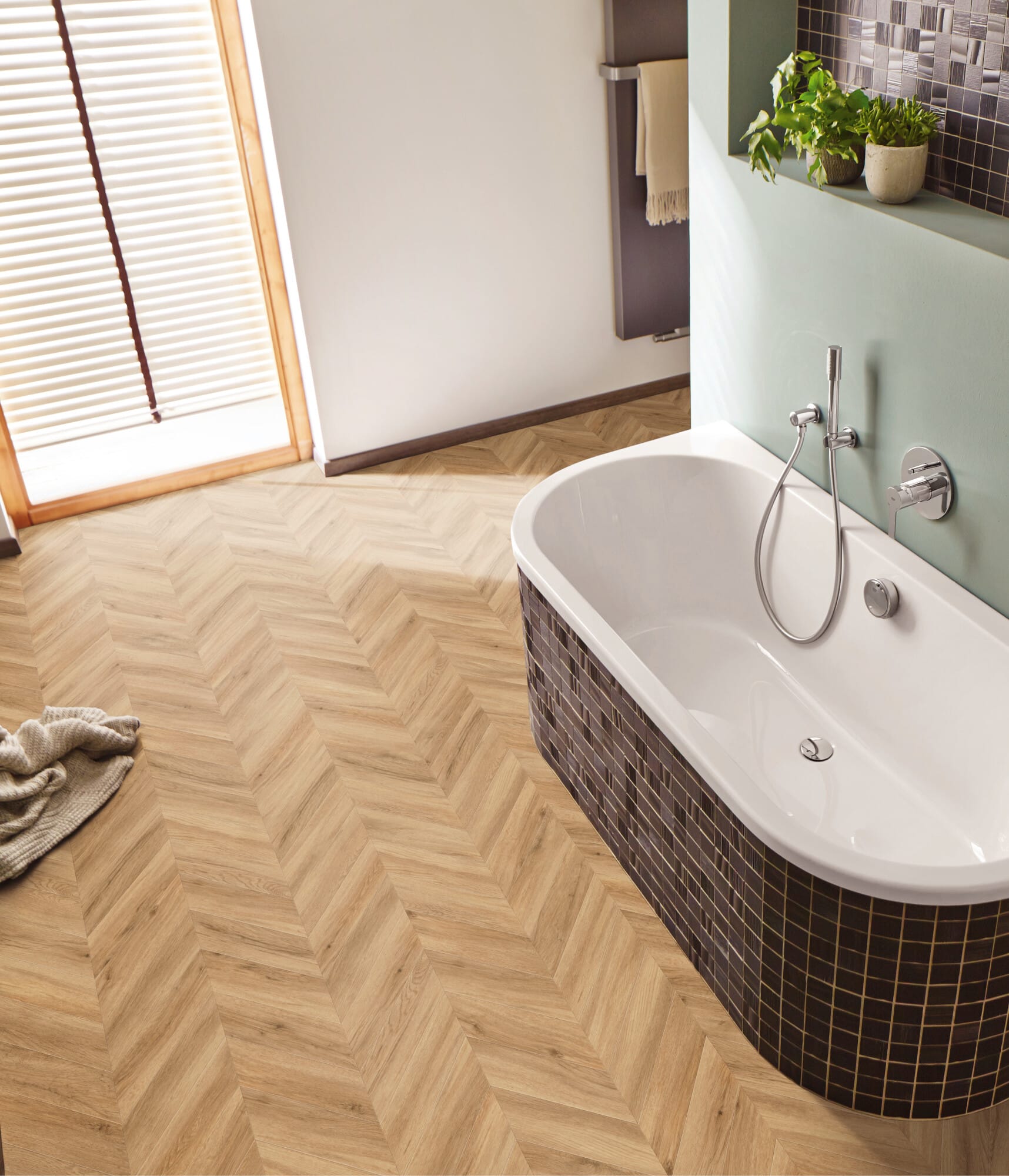 Ein modernes Badezimmer mit einer freistehenden Badewanne, einem hölzernen Fischgrätboden und einer sanft grünen Wand. Im Hintergrund sind Pflanzen und eine helle Fensterfront zu sehen.