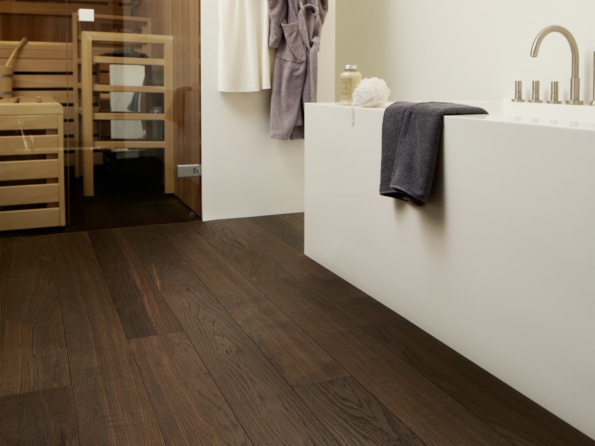 Ein modernes Badezimmer mit dunklem Holzboden, einer freistehenden Badewanne und einer Glaswand, die zu einer Sauna führt. Ein Handtuch und eine Duschschwamm liegen auf der Badewanne. Im Hintergrund sind Holzregale sichtbar.