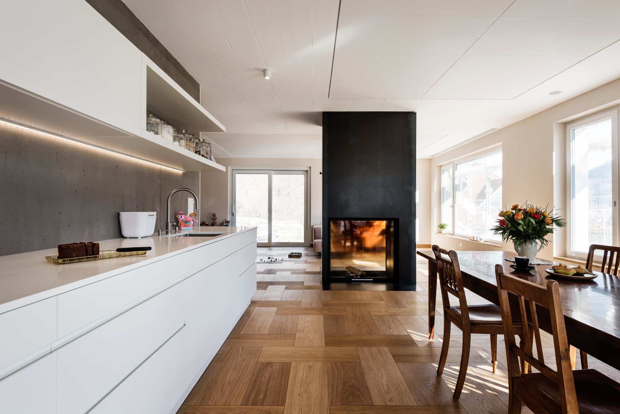 Moderne Küche mit weißen Schränken, einer zentralen Kochinsel und einem eleganten schwarzen Kamin. Große Fenster sorgen für viel Tageslicht, während ein Holztisch mit Stühlen und einer Blumenvase im Vordergrund eine einladende Atmosphäre schafft. Der Boden ist aus Holzparkett, das einen warmen Kontrast zu den hellen Wänden bietet.