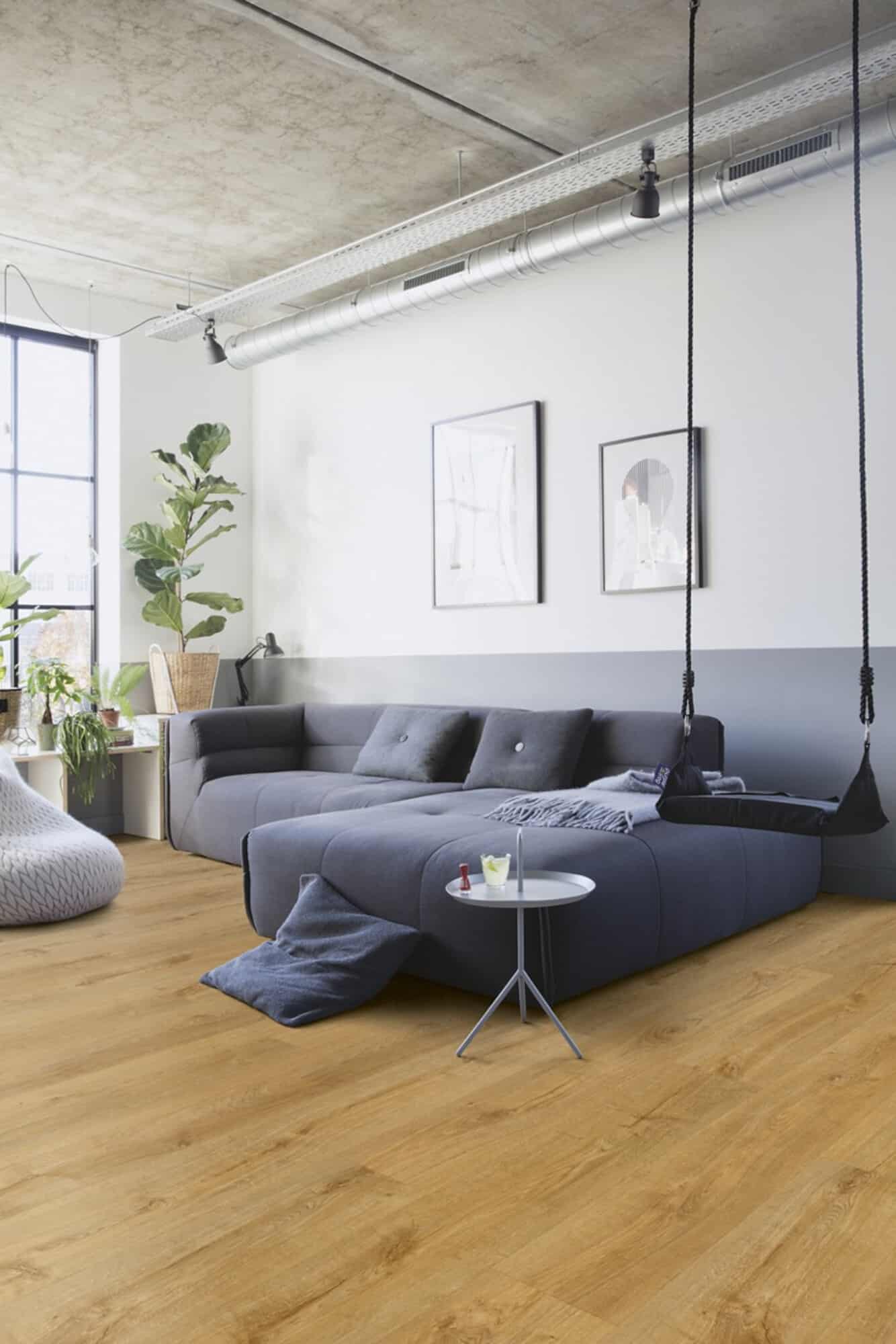 Ein modernes Wohnzimmer mit einem grauen Sofa, einem kleinen Beistelltisch und einem Hängesessel. Große Fenster lassen viel Licht herein, während Pflanzen für eine frische Atmosphäre sorgen. Der Boden ist aus hellem Holz.