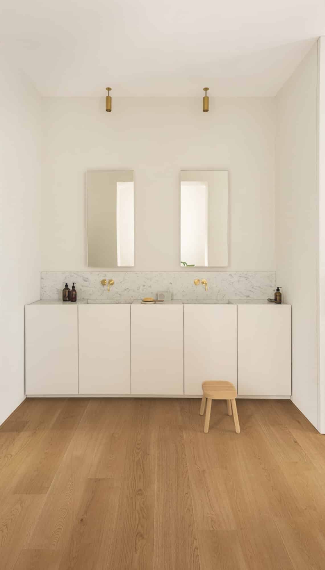 Ein modernes Badezimmer mit hellen Wänden, einem weißen Unterschrank und zwei rechteckigen Spiegeln über einem marmorierten Waschbecken. Es gibt goldene Wasserhähne und eine kleine Holzbank auf dem warmen Holzboden.