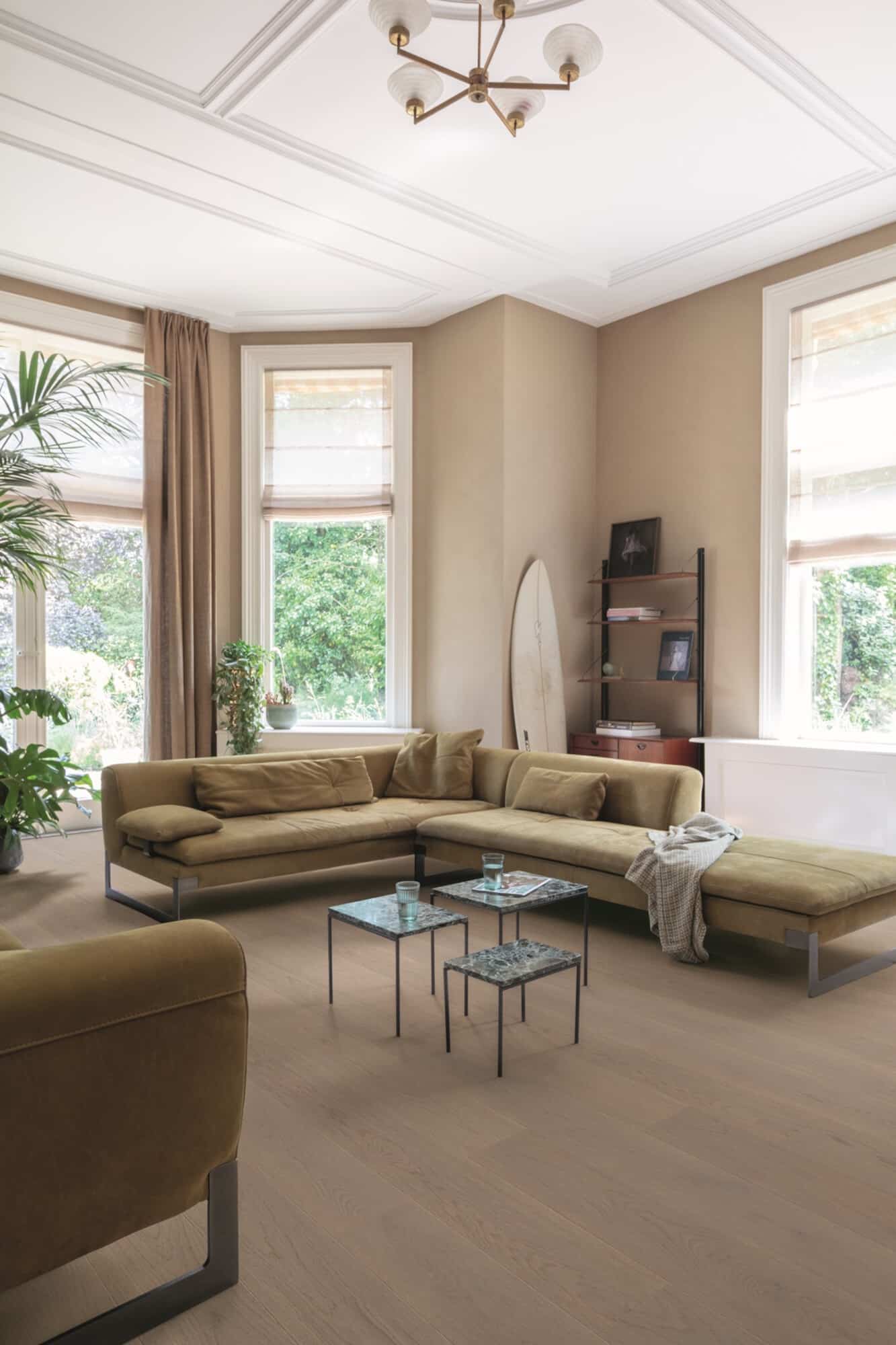 Heller, moderner Wohnraum mit einem beigen Sofa und gläsernen Couchtischen. Große Fenster lassen viel Tageslicht herein, während Pflanzen und ein Surfbrett für eine entspannte Atmosphäre sorgen. Die Wände sind in warmen Tönen gehalten und die Decke hat eine elegante Gestaltung.